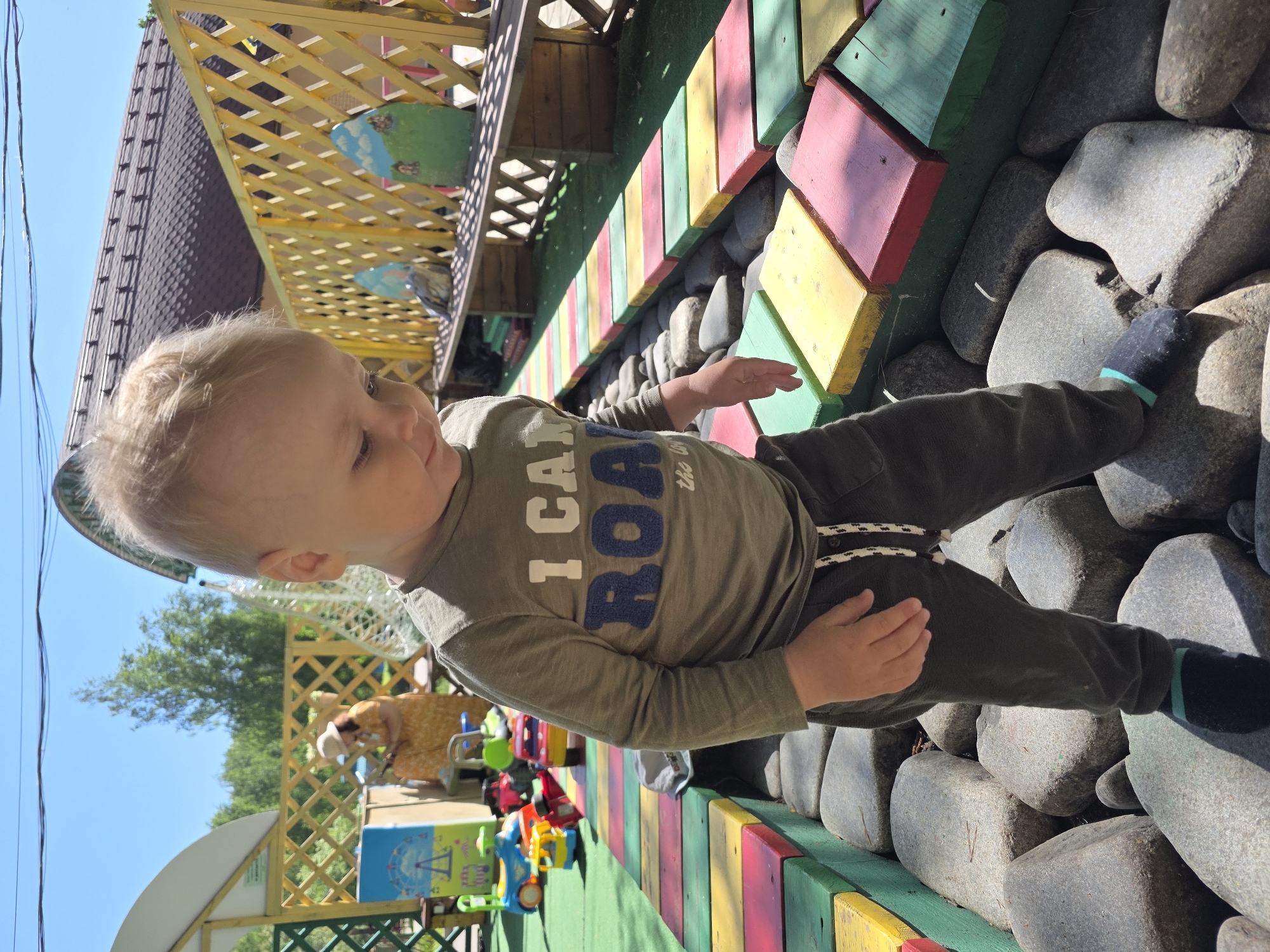 A young boy with fair hair stands on a stone wall next to a colorful walkway with green, yellow, and red wooden planks in what appears to be an outdoor recreational area. The boy wears a long-sleeved shirt with the words "I Can ROAR" on the front and dark-colored pants. His feet are clad in socks with teal accents. To the left, there is a green structure with a wooden lattice fence and a colorful display of toys. A building with a distinctive roofline is partially visible in the background. The location is Chaikovsky, Russia.