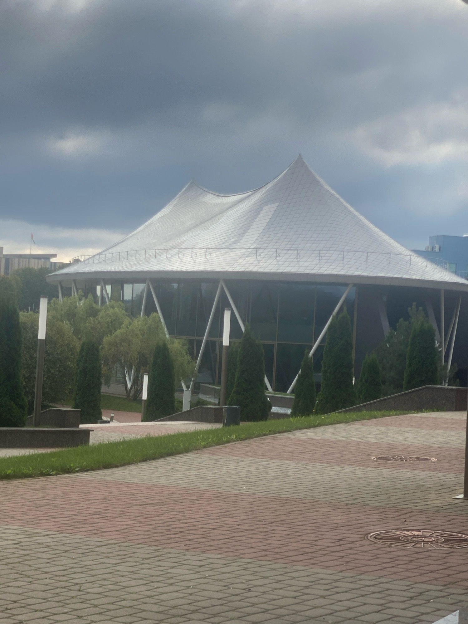 The image depicts the National Library of Belarus in Minsk, Belarus. The building features a distinctive diamond-shaped architectural design, with a large, multifaceted facade. A wide, paved walkway with a patterned brick surface extends in the foreground, leading towards the library. Tall, narrow evergreen trees and smaller shrubs are planted in intervals along the walkway and around the base of the building, interspersed with modern lampposts. The sky above is overcast with dark, heavy clouds, suggesting a gloomy or potentially stormy day. The overall scene is one of modern architecture set within a landscaped urban environment.