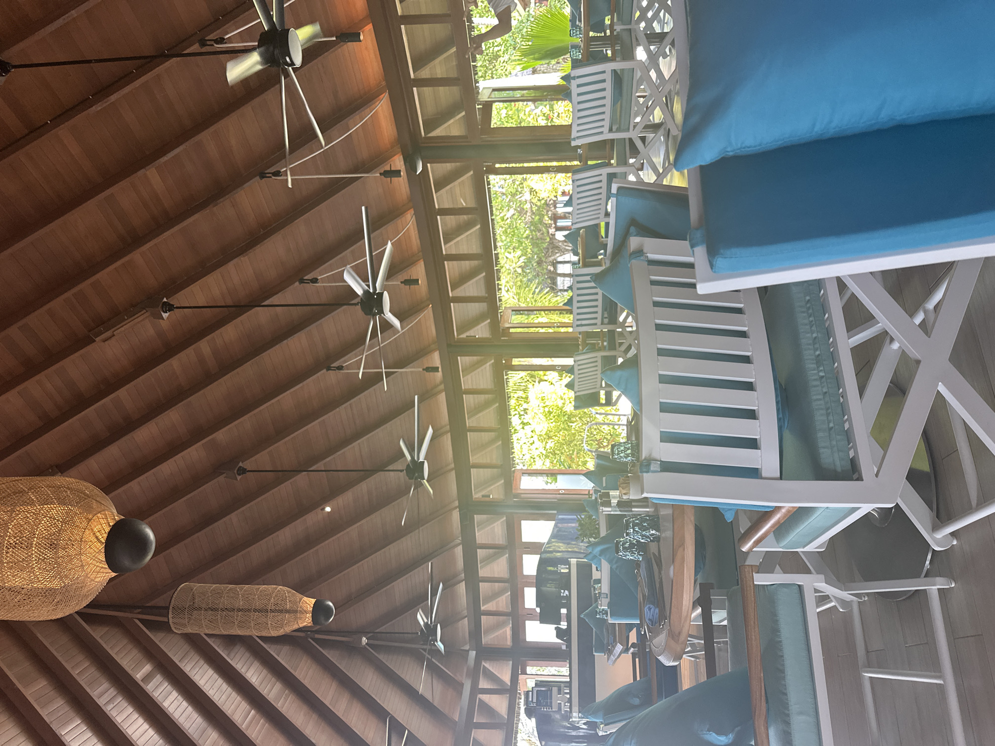 The image shows a wide shot of a restaurant interior, likely in the city of Halaveli, Maldives. The restaurant features a high, vaulted wooden ceiling with exposed beams, adorned with two large woven pendant lamps. Several ceiling fans are visible, suspended from the ceiling. The dining area is furnished with white tables and chairs, many of which are accented with bright turquoise cushions and pillows. The tables are set with glasses, plates, and napkins. Large windows or open doorways offer a view of lush green foliage and palm trees, suggesting an outdoor or tropical setting. The flooring appears to be light-colored wood or tile. In the background, a bar area is partially visible, with a counter and some equipment. There are no people clearly visible in the foreground, but there may be staff in the background. The overall atmosphere is one of tropical elegance and comfort.