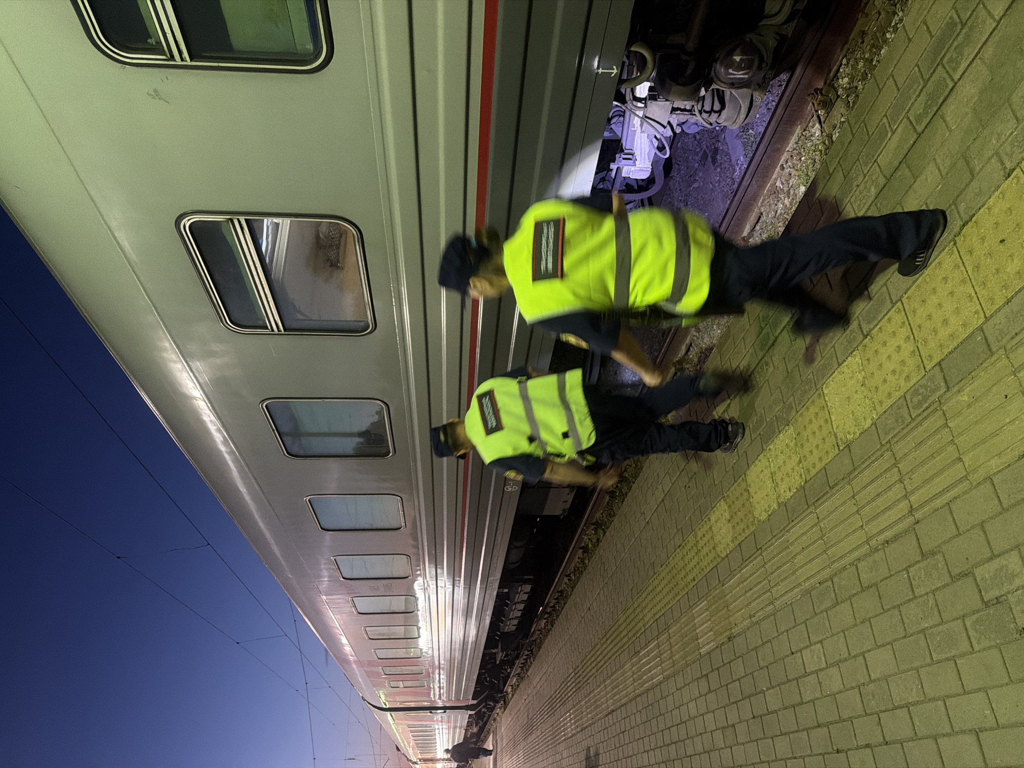 Two individuals wearing bright yellow safety vests and dark pants are walking on a tiled train platform at dusk. One individual is closer to the camera and slightly to the right, their face obscured by motion blur. The other individual is further away and to the left. They are walking alongside a long, grey train with multiple windows. The train appears to be stationary. The platform has tactile paving for visually impaired individuals, indicated by yellow textured tiles. The sky is a deep blue, suggesting it is either early morning or late evening. The environment appears to be a train station. The city of Balashov, Russia, is mentioned as the location of this scene.