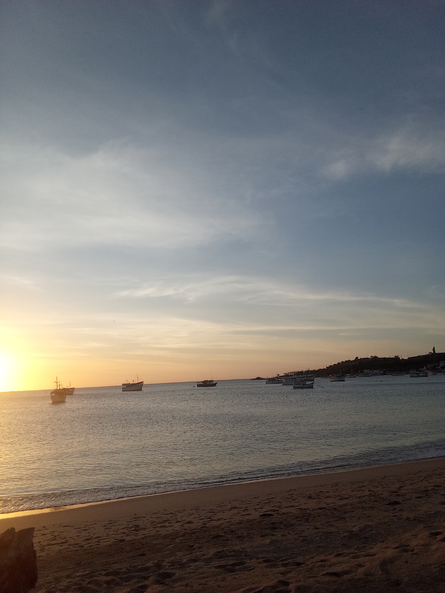 The image displays a coastal sunset scene in Juan Griego, Venezuela. The horizon line bisects the frame horizontally. The left side features a low, intense orange and yellow glow, indicating the setting sun, which casts warm light across the water's surface. The sky transitions to light blue and greyish-blue towards the right, with faint, wispy clouds present throughout the upper two-thirds of the image.
The midground is dominated by a calm body of water with gentle ripples. Approximately 15 boats of varying sizes are visible on the water. Three larger vessels with discernible superstructures or masts are silhouetted against the brighter light on the left. Numerous smaller, lighter-colored boats are scattered closer to the distant right shoreline.
The foreground consists of a dark, sandy beach with visible footprints. A line of white foam marks the gentle waves breaking at the water's edge. A dark, indistinct object is partially visible in the bottom-left corner. The background on the right reveals a dark, undulating landmass, likely a hillside or coastline, with faint, indistinct structures, including a potential tower or lighthouse, visible on its crest. No individuals or specific interactions are discernible within the frame.