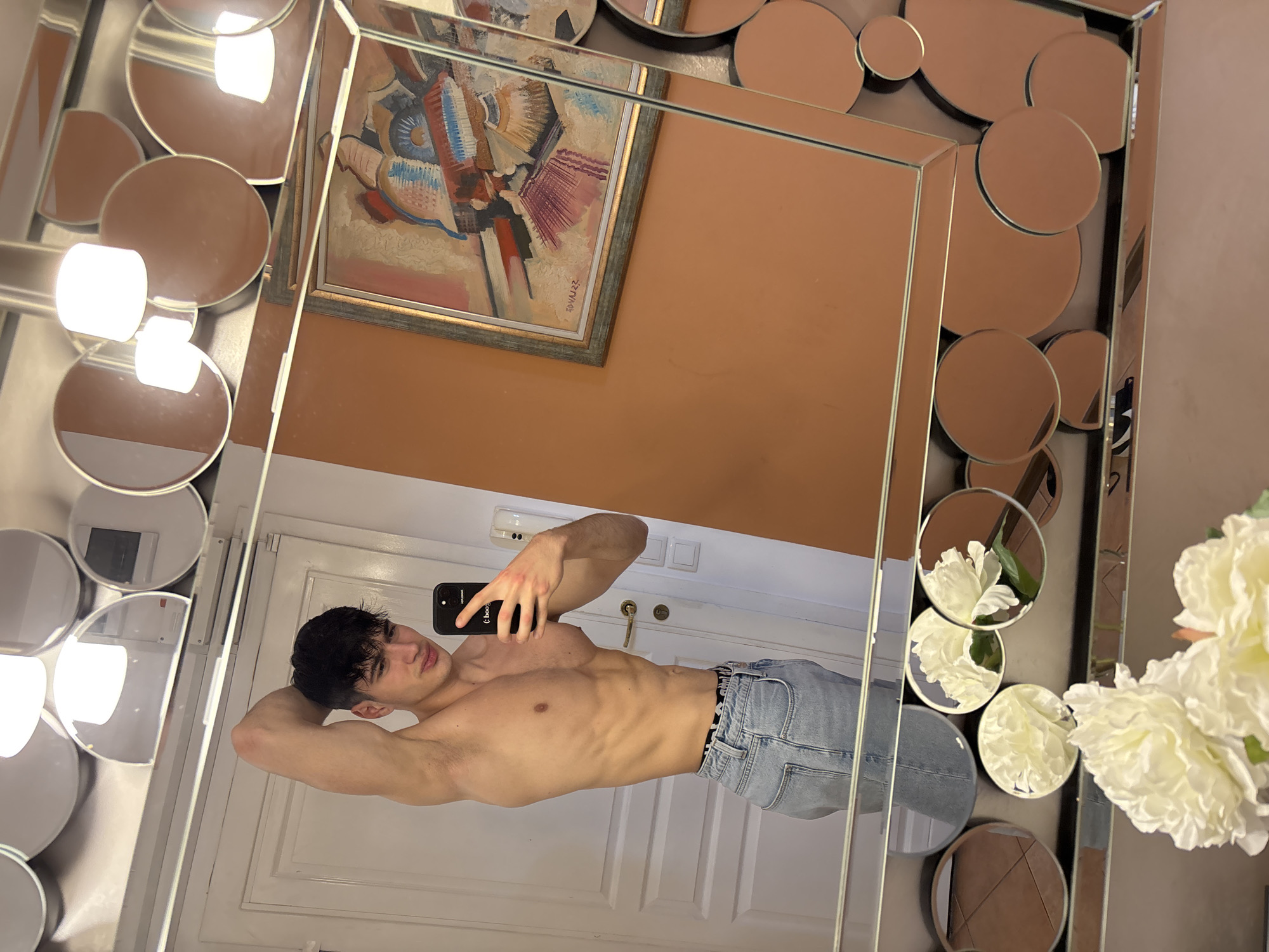 A young man takes a mirror selfie indoors in Plovdiv, Bulgaria. He is shirtless, displaying a muscular physique, and wears light blue denim jeans with a visible "CALVIN KLEIN" waistband. He holds a black smartphone with a partially visible "boo." logo to capture his reflection, posing with his right arm bent behind his head and a neutral expression.
The large, ornate mirror consists of a central rectangular panel surrounded by numerous smaller circular and semi-circular mirrors. These smaller mirrored sections reflect elements of the room, including bright light sources and a vase holding white flowers on the right side.
The background reflected in the main mirror includes a wall with white double doors or panelling. Above the man, a section of an orange-brown wall displays a framed, abstract or cubist painting featuring vibrant colors like blue, red, and brown. Text "20 VALEZ?" is visible on the painting. A light switch or outlet is also discernible on the wall behind his head in the reflection. The scene is illuminated by artificial indoor lighting.