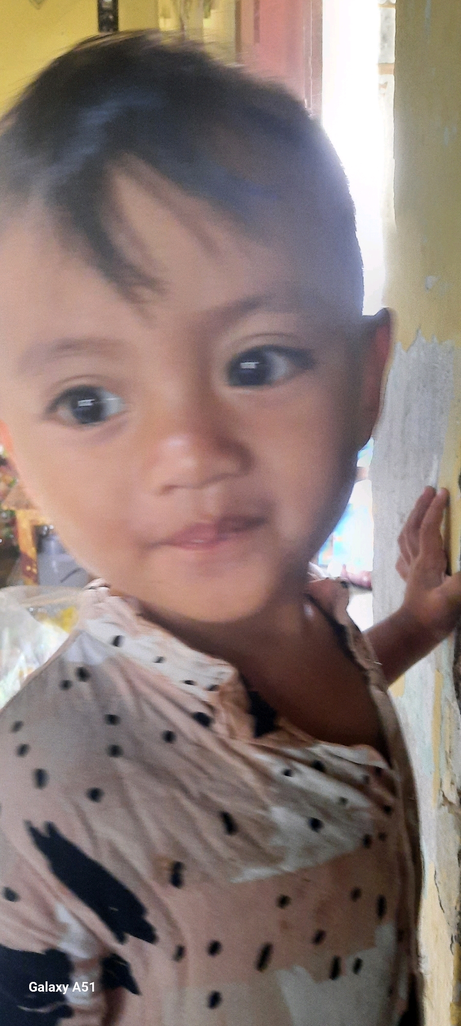 The image is a close-up, vertical, slightly blurry photograph of a young child in Jatitengah, Indonesia. The child, with dark hair and brown eyes, is looking directly at the camera. They are wearing a light-colored shirt with dark polka dots. The child is leaning against a wall that appears to be peeling or textured. In the background, there is a hint of a doorway and some indistinct objects. The overall lighting suggests an indoor or shaded outdoor environment. The bottom left corner of the image displays "Galaxy A51".