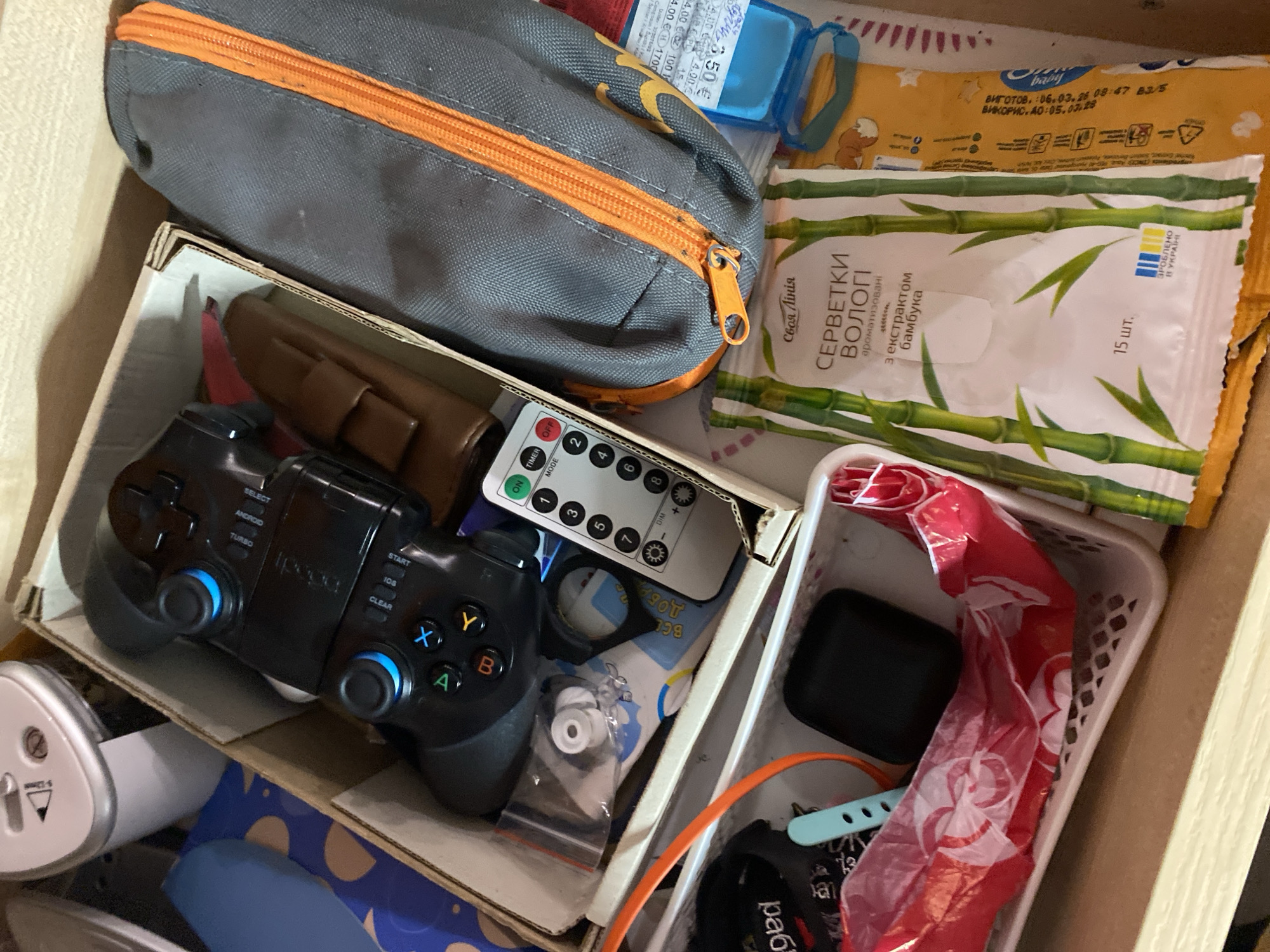 The image shows a close-up, overhead view of a drawer filled with various items. In the center of the frame, a black game controller with blue accents is visible inside a cardboard box. To the right of the box is a grey pencil case with an orange zipper. Below the pencil case and partially obscuring the box is a package of wet wipes with bamboo imagery and Ukrainian text. To the left of the game controller, a white plastic basket contains a red and white plastic bag, a black square object, and a blue wristband. Further to the left, a white electric pencil sharpener is partially visible. The overall impression is of a cluttered drawer containing personal electronics and stationery. The specific location in Kryvyi Rih, Ukraine, cannot be determined from the visual information provided.