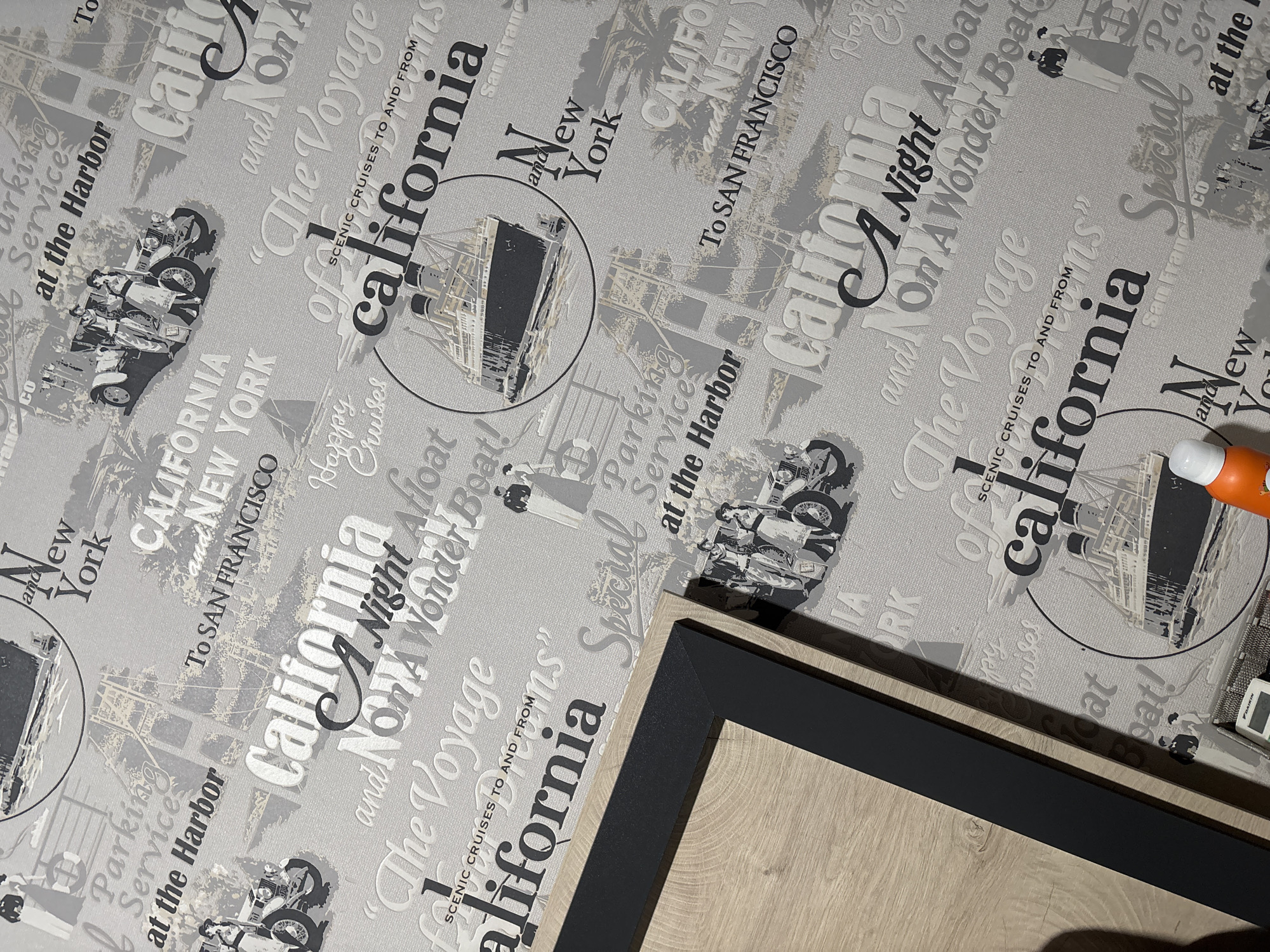 The image displays a section of a wall covered in patterned wallpaper. The wallpaper features a vintage travel theme with various text and illustrations, including ships, vintage cars, and stylized fonts. Prominent text includes "California," "New York," and "San Francisco," along with phrases like "Parking Service at the Harbor" and "The Voyage of California." There are also illustrations of people in vintage attire. In the lower left quadrant, a corner of a light-colored wooden surface with a dark frame is visible, suggesting it's part of a piece of furniture or decor. A white bottle with an orange dispenser is also partially visible at the bottom of the frame. The scene appears to be an indoor setting. The provided coordinates do not relate to Varna, Bulgaria, nor do they provide any geographical context.