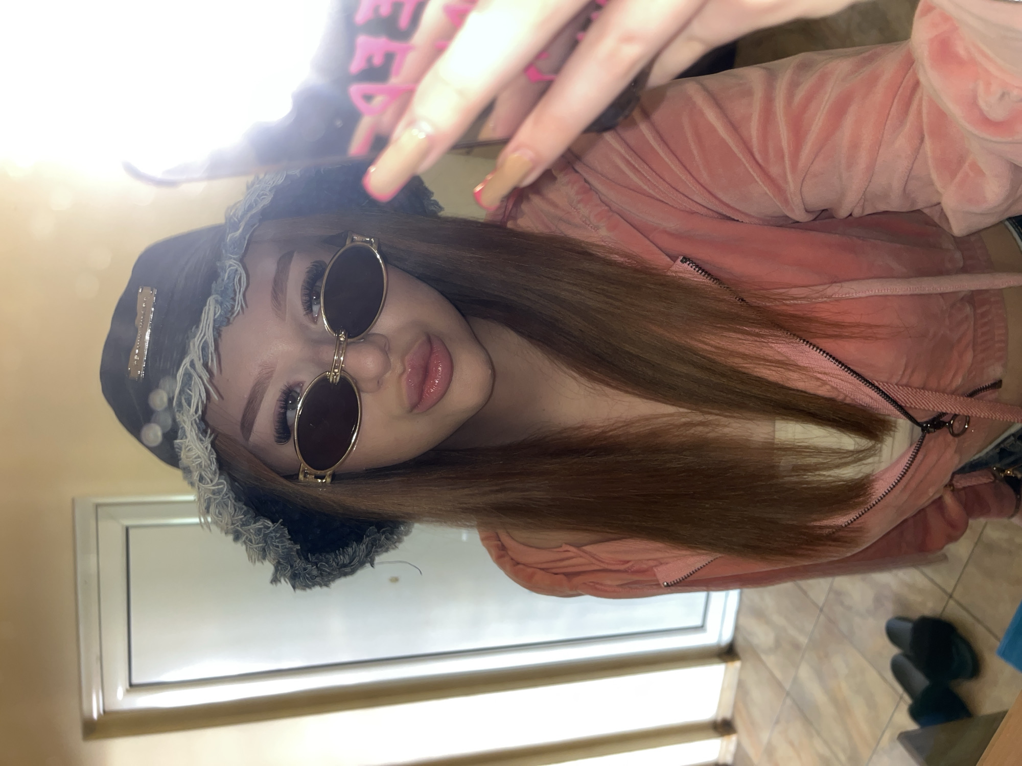 A young woman is seen in a selfie, wearing sunglasses, a black hat with a frayed trim, and a light pink velvet jacket. She has long, light brown hair. Her fingers are visible in the top of the frame, with long nails, some painted pink. The background includes a white door or window frame. On the floor, there's a pair of dark-colored slippers. The location details of the city of Varna, Bulgaria, cannot be determined from this single image.