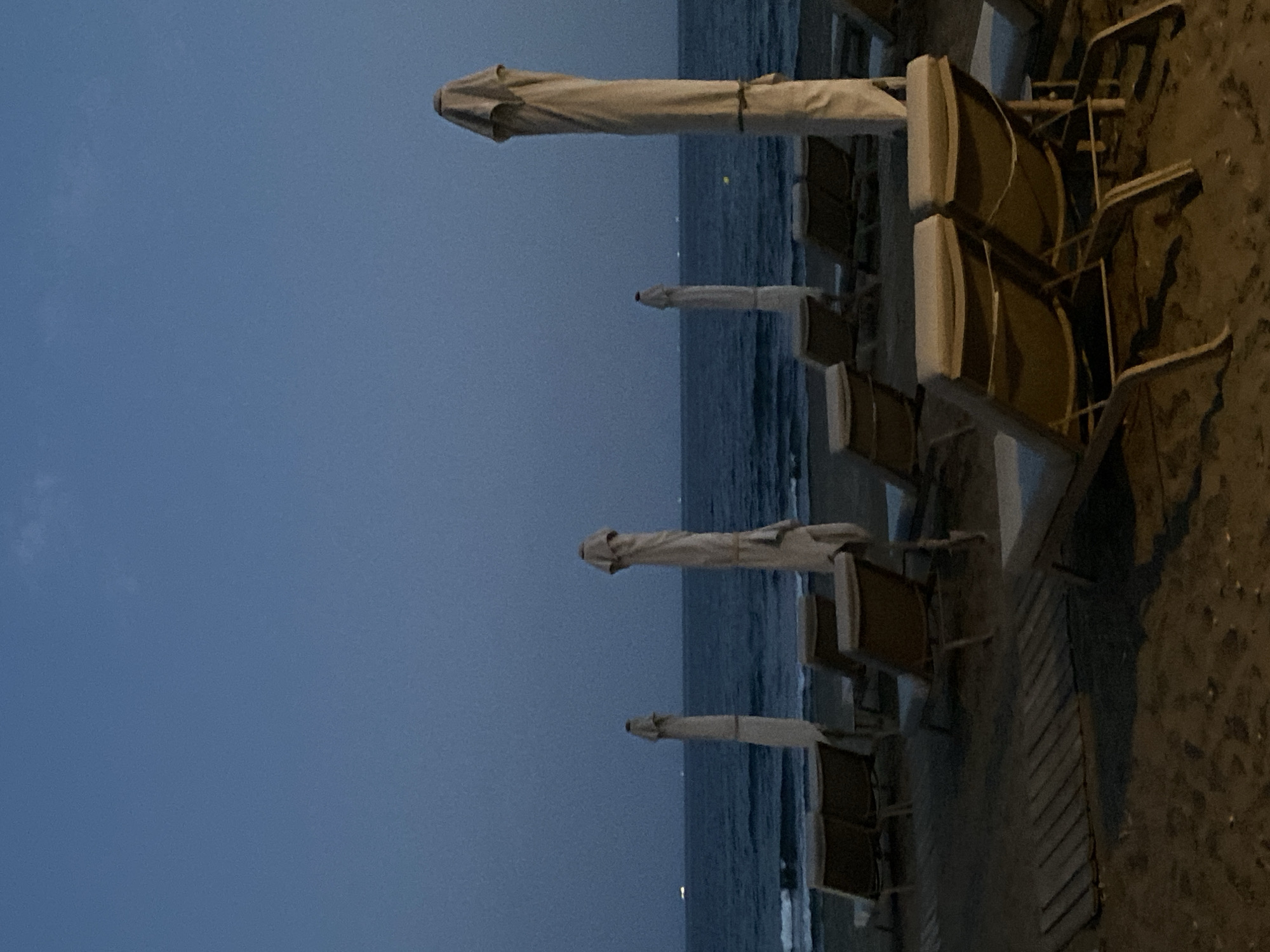 A nighttime view of a beach in Estepona, Spain shows several empty lounge chairs and closed umbrellas on the sand, facing a calm sea. The sky is a deep twilight blue, with a few distant lights visible on the horizon, possibly from boats or the coastline. The sand is dark and slightly damp, with a wooden walkway visible in the foreground. The overall scene conveys a sense of stillness and quietude, as if the beach has been deserted for the night.