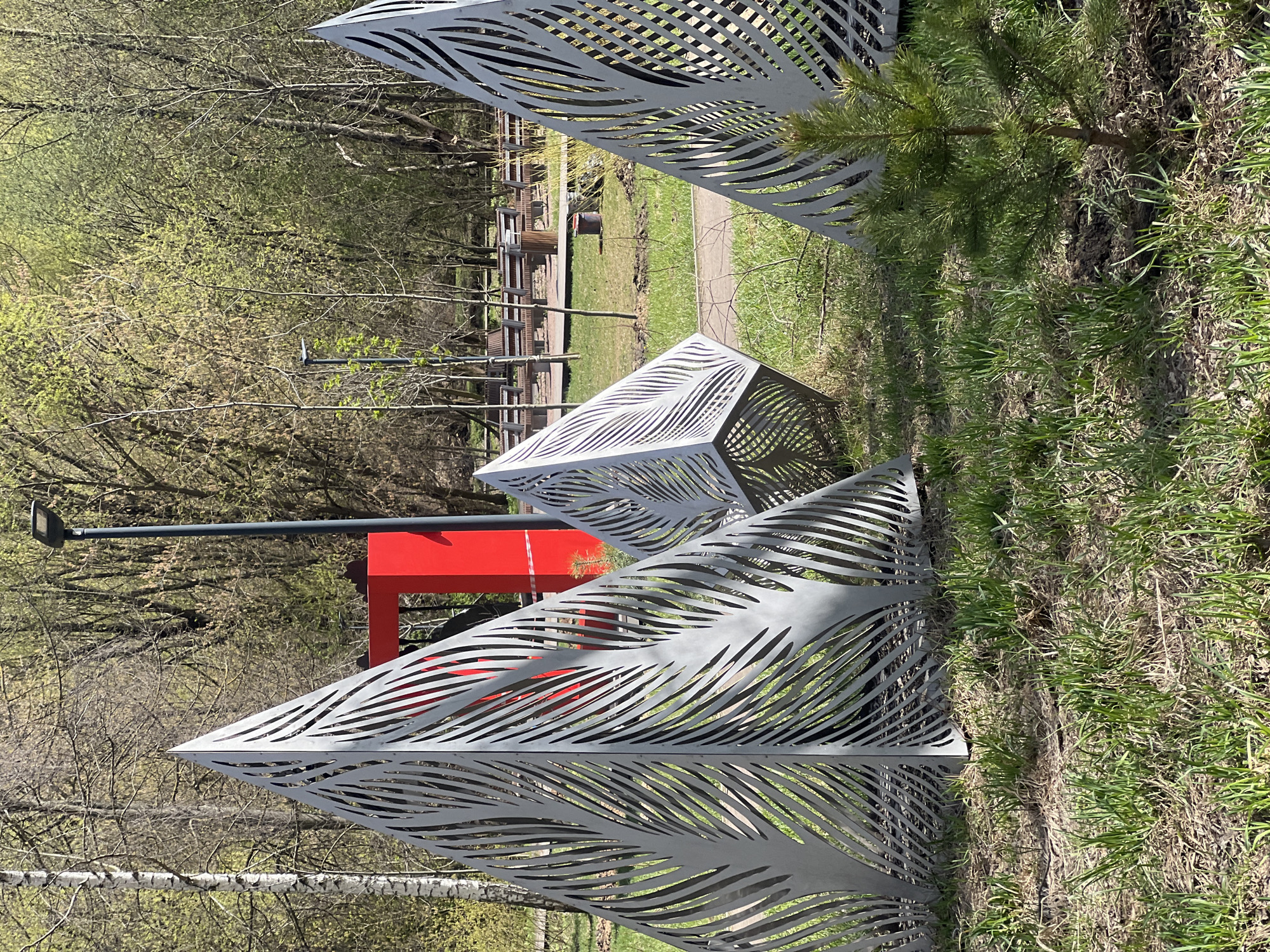 The image captures a peaceful outdoor scene in a park in Khimki, Russia, dominated by several modern art installations. The setting is likely early spring, as many trees in the background are still bare-branched, but some show the first delicate signs of light green foliage. The ground is covered with a mix of green and dry grass.
In the foreground and midground, three distinct geometric art pieces are visible. These structures are made of a light-colored, possibly metallic material, intricately perforated with wavy, organic, or leaf-like patterns. One large, triangular prism-like structure is prominent on the left, while a smaller, cube-shaped piece with similar patterns is centered behind it. A third, partial triangular structure is seen on the right, partially obscured by a small, vibrant green pine tree planted in the grass.
Further into the midground, a bright red, rectangular structure, possibly a part of a playground or an abstract bench, stands out against the muted greens and browns of the natural environment. To its left, a tall, dark grey street lamp with a distinct light fixture rises towards the sky. In the deeper background, a paved path meanders through the park, and several simple wooden benches and tables are scattered along it.
The scene is illuminated by bright daylight, casting clear shadows, suggesting a sunny day. No people are visible, and no specific activity is taking place, creating a serene and quiet atmosphere. There is no visible text in the image.