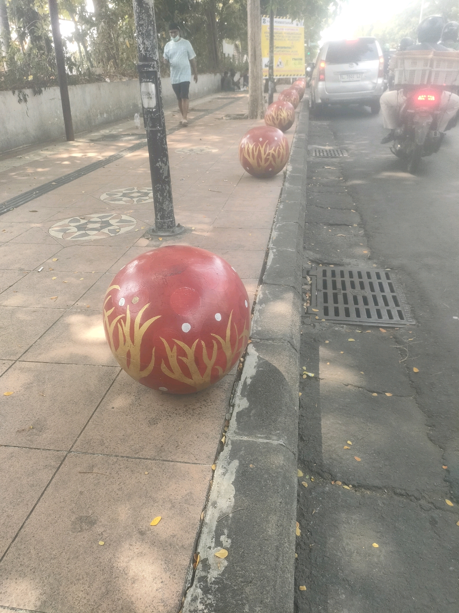 This image depicts a street scene in Surabaya, Indonesia, focusing on a sidewalk adorned with decorative red spheres. The foreground is dominated by a large, red sphere with gold leaf-like designs and white dots, placed on a paved sidewalk. To the right, a street with a grey concrete curb is visible, featuring a metal drain cover. A line of similar red spheres recedes into the distance along the sidewalk. In the background, a person wearing a light blue shirt and dark shorts walks on the sidewalk, and a silver car and a motorcycle with a rider are on the street. Trees and foliage line the far side of the sidewalk, partially obscuring a wall and some signage. The overall scene suggests a sunny day in an urban environment.
