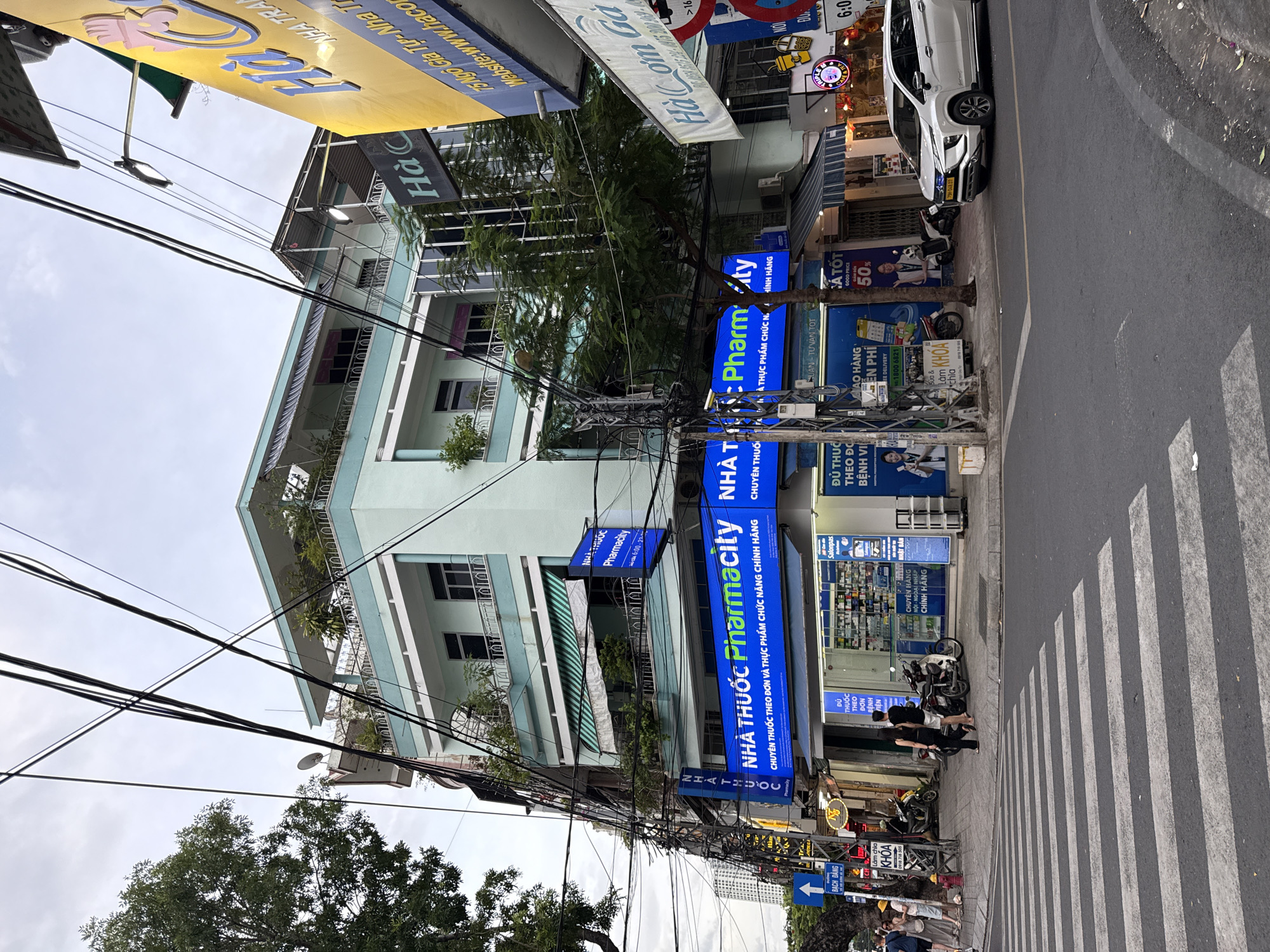 The image shows a street scene in Nha Trang, Vietnam, with a prominent Pharmacity drugstore on the corner of a light blue, multi-story building. The drugstore has a bright blue illuminated sign that reads "NHÀ THUỐC Pharmacity" and a smaller sign below it in Vietnamese. There are awnings over the windows, and various signs advertising products and services are visible, including one for "GOOD PRICE" and "50%". Several motorcycles are parked outside the drugstore, and a white SUV is driving on the street. A pedestrian crossing is in the foreground, and a group of people are walking on the sidewalk to the left. Overhead, a tangled network of electrical wires crisscrosses the sky. In the background, other buildings and a distant cityscape are visible. A yellow sign for "Ha Co" is visible to the right of the Pharmacity, and a sign for "Ha Com Ga" is also visible.