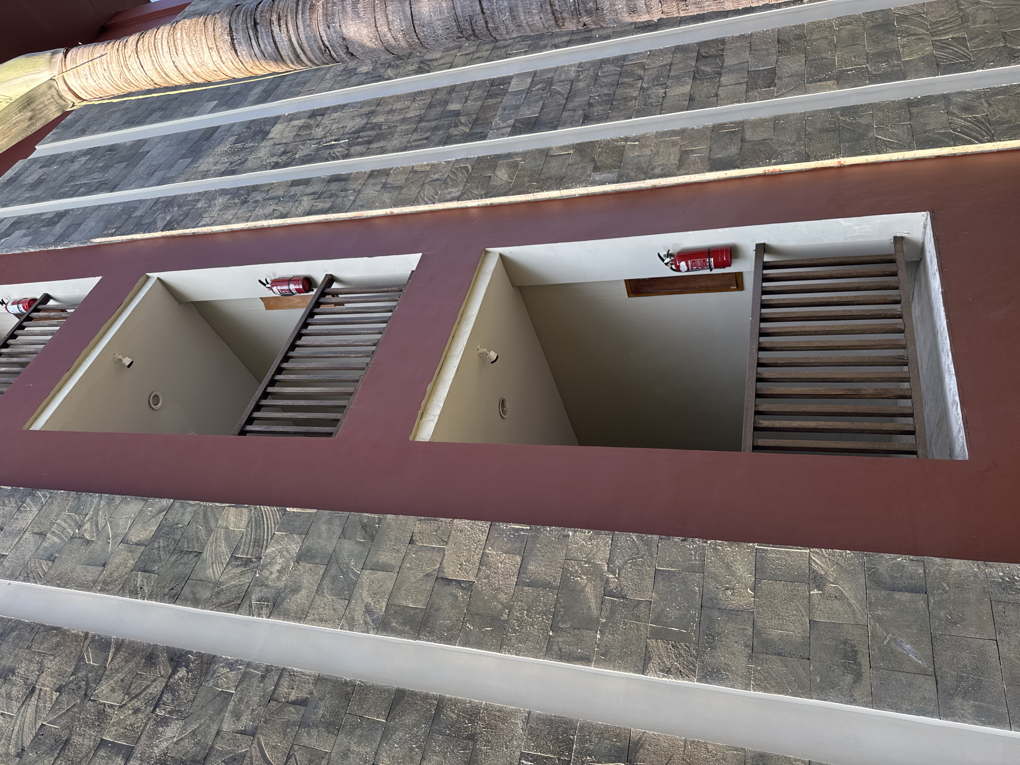 The image presents an upward angle view of a building exterior, possibly a hotel or resort, located in Kuta Selatan, Indonesia, characterized by distinct architectural elements.
The building features a prominent reddish-brown horizontal band, possibly a facade or wall. Within this band are regularly spaced, recessed openings. Each opening reveals a stark white interior space with an overhead light fixture or camera present. A series of horizontal wooden slats, arranged vertically, are also seen in front of the opening.
Mounted above each of these openings is a red fire extinguisher. The building is constructed from a patterned stone or brick material, with alternating rows of dark and light colored stones.
Above the building, a fragment of a cylindrical structure, possibly a palm tree trunk or some other type of architectural design, is visible. The overall impression is one of a modern architectural design, built with attention to material contrasts and visual rhythm.
