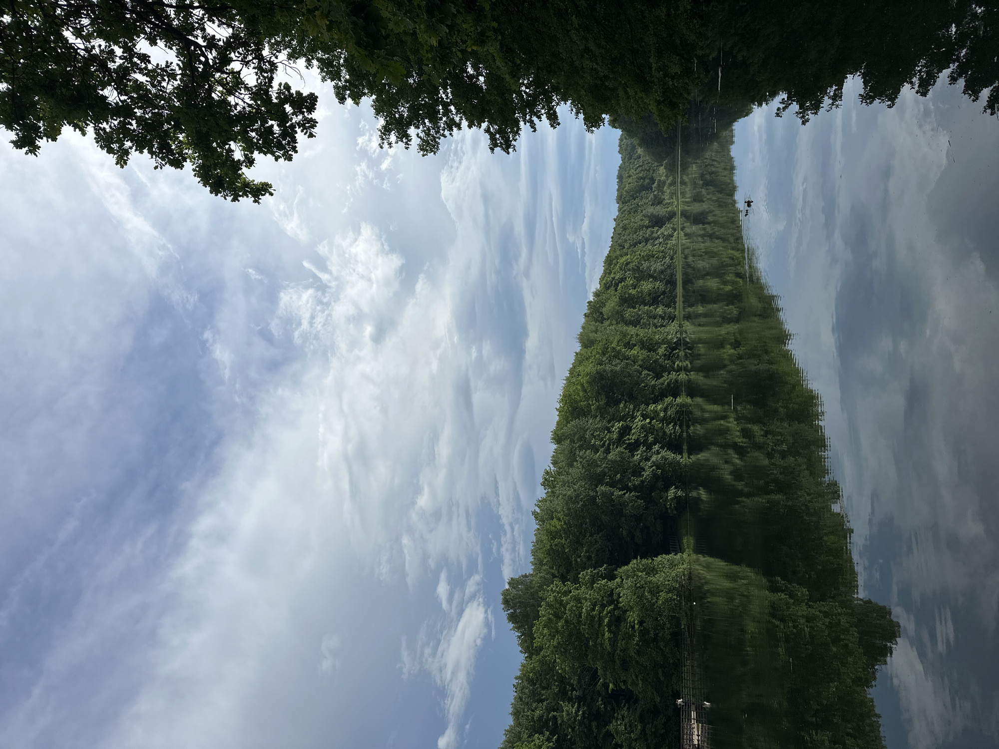 The image portrays a serene natural landscape, oriented as if viewed horizontally, featuring a long, calm body of water, likely a river or canal, extending into the distance. Both banks are densely lined with lush green trees, creating a verdant corridor. Above the treeline, a partly cloudy sky with white and grey clouds against a light blue backdrop is visible. The tranquil water perfectly mirrors the sky and the trees along the banks, producing a striking and clear reflection that creates an almost symmetrical composition.
In the middle distance of the water, a small, dark-colored bird, possibly a duck or coot, is swimming. The clarity of the reflection is a prominent feature, almost duplicating the real landscape above the waterline. The lighting suggests it is daytime, likely late morning or early afternoon, with ample light illuminating the scene. No people are visible, nor is any discernible text present. The overall setting conveys a sense of peace and natural beauty, consistent with a scenic waterway in Nizhny Novgorod, Russia.