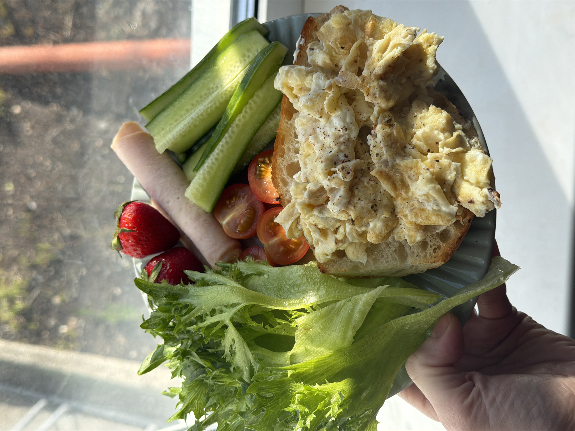 A plate of food is being held up in front of a window. The plate contains a slice of bread topped with scrambled eggs, alongside slices of cucumber, halved cherry tomatoes, rolled-up ham, two strawberries, and lettuce. The setting appears to be indoors, near a window. There is no evidence to confirm if this image was taken in Perm, Russia. No people other than a partial view of the arm and hand holding the plate are visible.