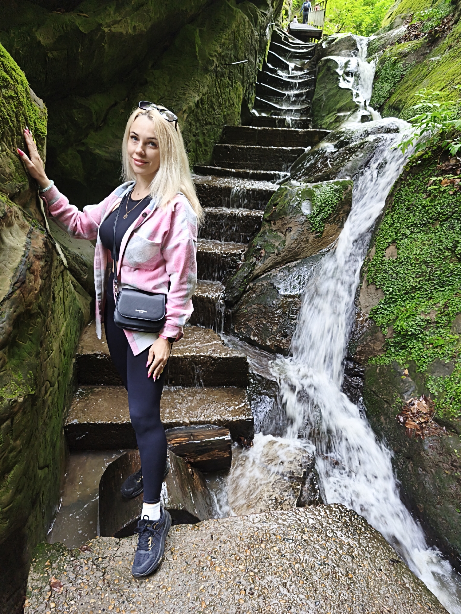 A fair-skinned woman with long blonde hair, wearing a pink flannel shirt over a black top, black leggings, and athletic shoes with white socks, stands on stone steps beside a waterfall. She also carries a black shoulder bag and wears sunglasses perched on her head. She touches a moss-covered rock face to her left. The waterfall cascades down the stone steps, which appear to be built into the natural rock formation. Moss and greenery cover the surrounding rocks, suggesting a damp, natural environment. Another person is visible on the stairs above. The location is Dandropov Cascade in Goryachiy Klyuch, Russia.