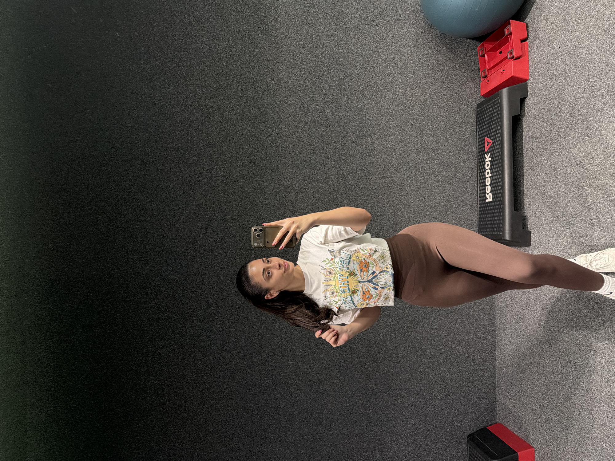 A woman is taking a selfie in a gym. She is wearing a white printed t-shirt and brown leggings, and white sneakers can be seen at the edge of the frame. The gym appears to have a grey floor and is equipped with a Reebok step platform and an exercise ball. The location is in Sofia, Bulgaria.