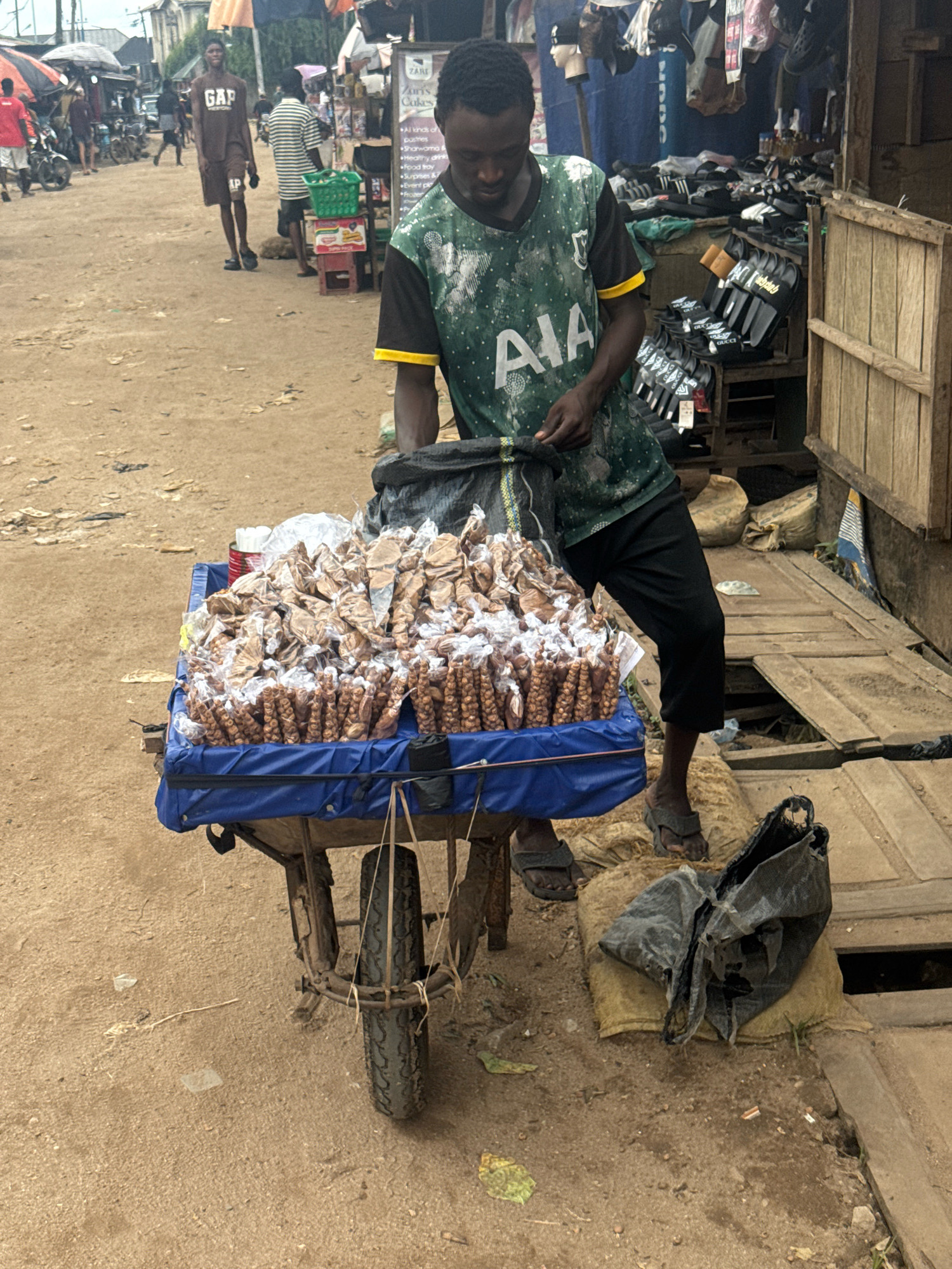 In the city of Ihiagwa, Nigeria, a vendor stands beside a wheelbarrow laden with goods. The wheelbarrow, draped in a blue tarp, holds numerous small, individually packaged food items, likely snacks, piled high. The vendor, a Black male wearing a green and white patterned shirt with yellow trim and dark pants, is in the process of handling a black plastic bag, his hands near the contents of the wheelbarrow. He wears dark sandals. The ground around him is dusty, dirt road, typical of an outdoor market setting. In the background, other market stalls and individuals are visible, with various goods displayed and people moving about. The overall scene depicts a busy marketplace environment.