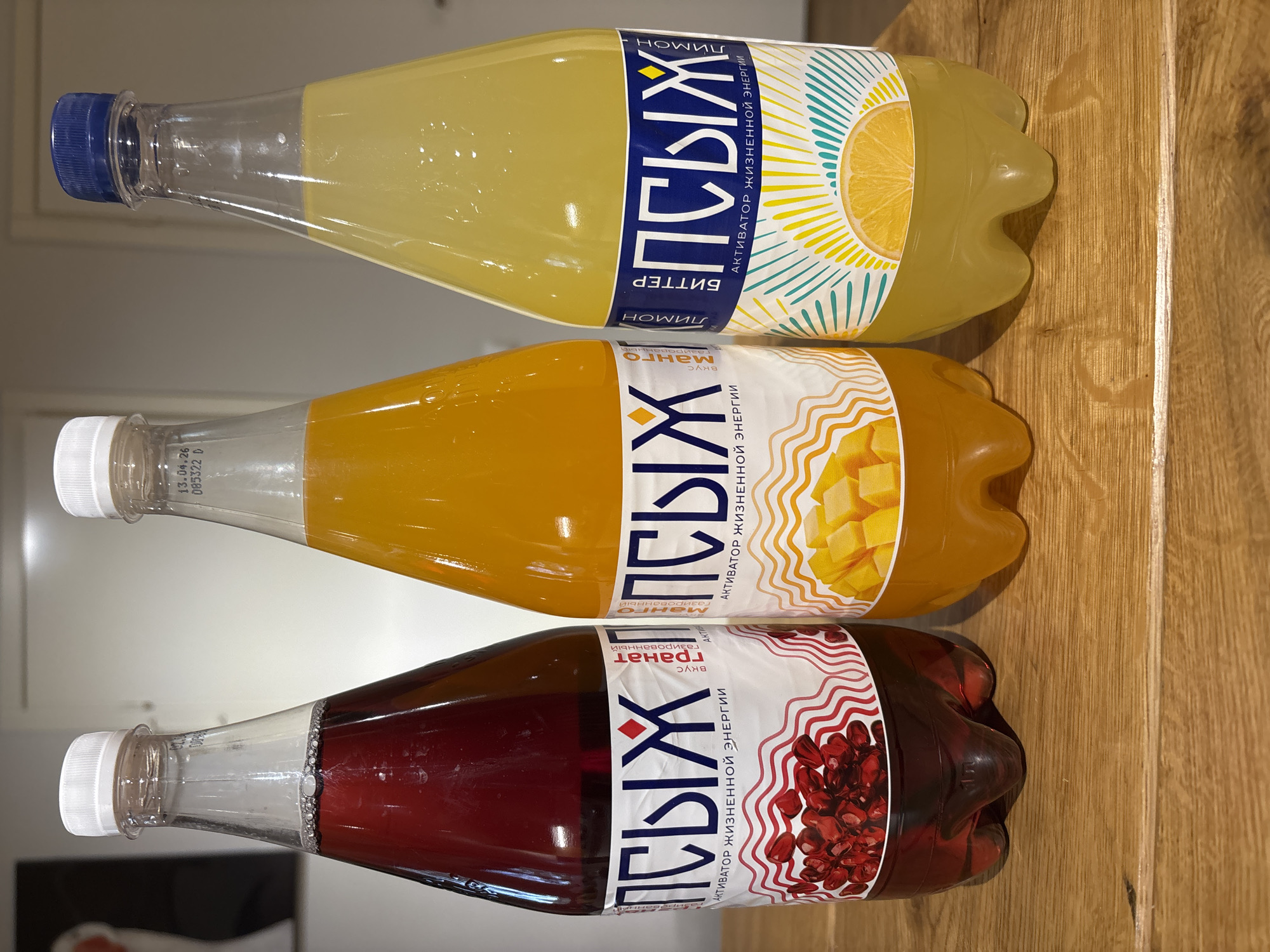 The image shows three plastic bottles of "PSYCH" (ПСЫХ) brand sparkling juice resting horizontally on a wooden surface. The top bottle contains a yellow liquid, labeled as "Bitter Lemon" (Лимон). The middle bottle holds an orange liquid, labeled as "Mango" (Манго). The bottom bottle contains a dark red liquid, labeled as "Pomegranate" (Гранат). The label text is in Russian, stating "Activator of Life Energy" (Активатор Жизненной Энергии). The bottles appear to be produced in Chaikovsky, Russia, based on the presence of Russian text and the brand name.