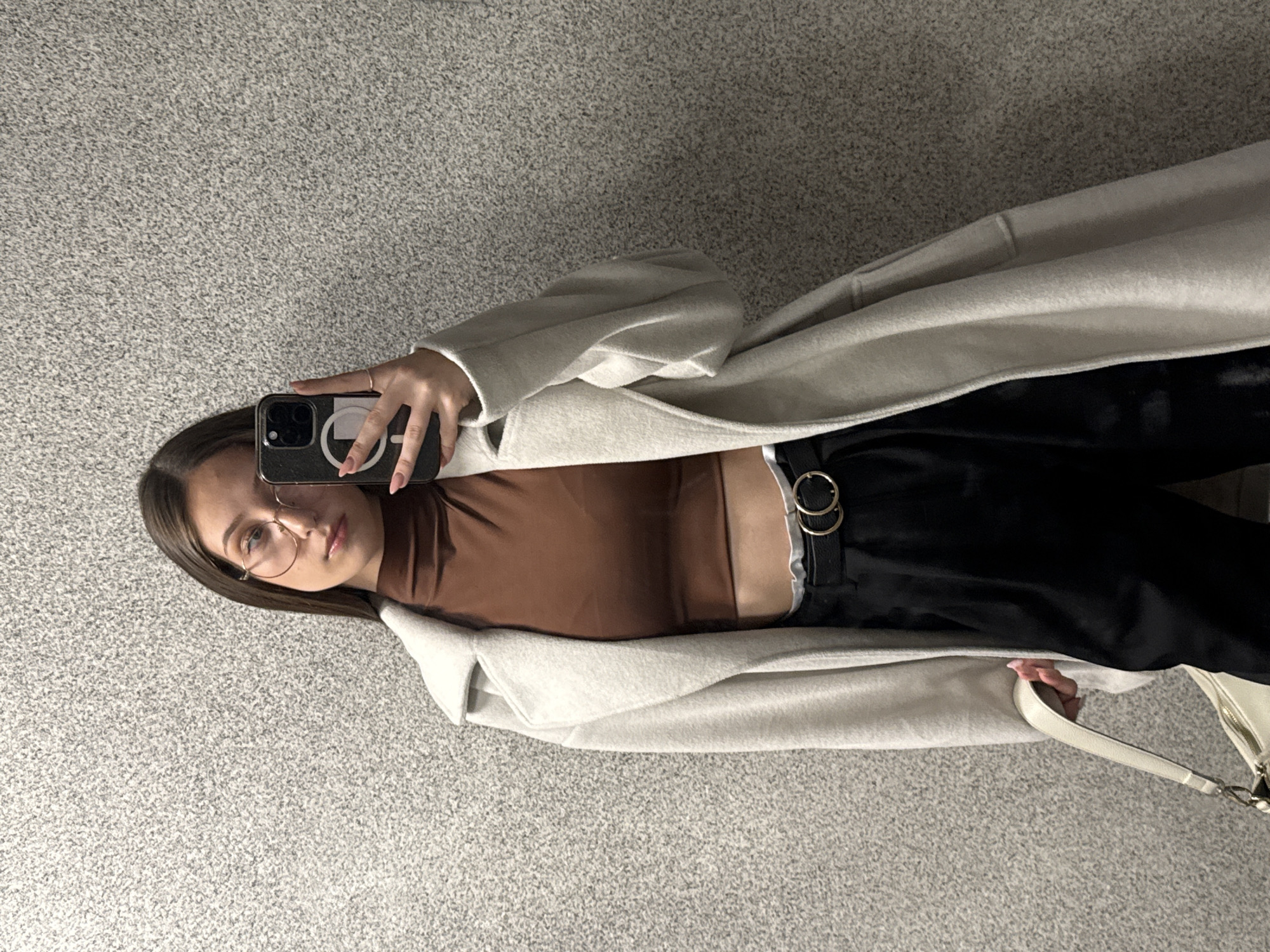 A young woman is captured in a mirror selfie, holding a black smartphone with a distinctive white circular logo on its case in her right hand. She has dark hair, is wearing round, gold-rimmed glasses, and looks directly towards the camera with a neutral expression.
She is dressed in a light-colored, possibly beige or off-white, long coat or cardigan that drapes open. Beneath the coat, she wears a fitted brown, possibly turtleneck or mock-neck, top. Her lower attire consists of black pants cinched with a black belt featuring two prominent interlocking gold-colored circular buckles. A white handbag is visible towards the bottom right, held in her left hand. A subtle ring is visible on a finger of her right hand.
The setting appears to be indoors, against a speckled gray and light beige textured surface, resembling carpet or a tiled floor, which suggests a consistent, possibly public or commercial, indoor environment. There are no direct textual cues other than the phone case logo and belt buckles. The location context provided is Sofia, Bulgaria.