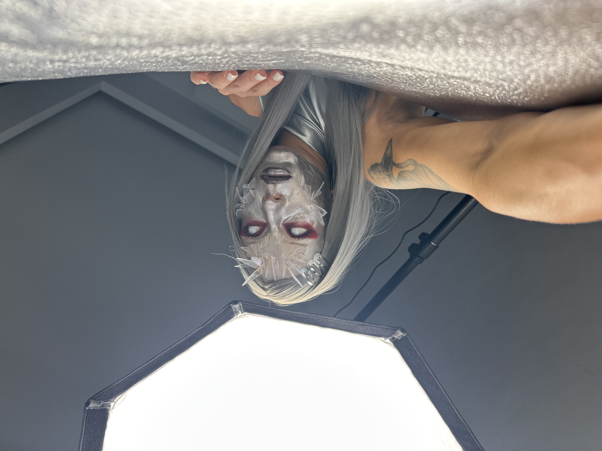 A low-angle, close-up, waist-up shot shows a person with pale grey hair, heavily stylized makeup, and a futuristic costume. The makeup includes silver face paint, white and red eyeshadow, and dark lipstick. Clear, jagged plastic pieces are adhered to the forehead and cheeks, resembling shards of ice. The person's eyes are wide open with red accents around them. They are wearing a silver, shiny garment. The person is leaning towards the right side of the frame, with their right hand resting on a textured, reflective surface that appears to be a white, crinkled fabric or backdrop. A tattoo of a bird is visible on their left upper arm. To the left of the frame is a large, illuminated softbox, a piece of photography lighting equipment. The background is a dark grey wall with architectural elements visible, suggesting an indoor studio setting. There are no identifiable landmarks or elements that suggest a specific location in North Kuta, Indonesia.