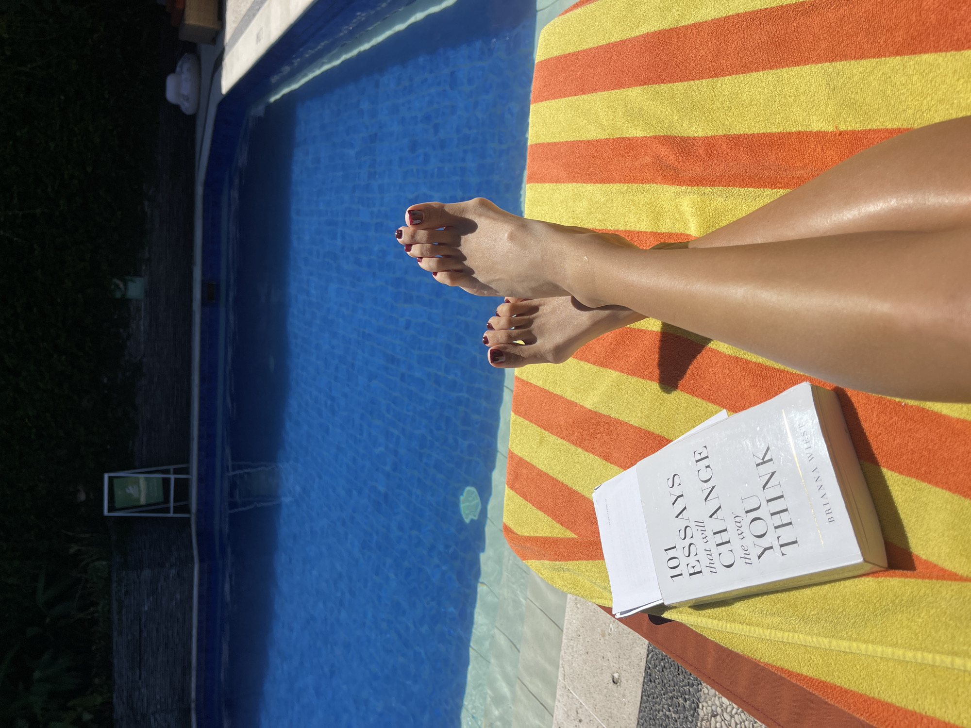 The image depicts a scene by a swimming pool in Kuta, Indonesia. A pair of tanned legs with red-painted toenails are positioned on a yellow and orange striped towel. The legs are crossed at the ankles, with a book resting near them. The book's title is "101 Essays That Will Change The Way You Think" by Brianna Wiest. The pool has blue tiled walls and clear water. Green vegetation borders the pool area.