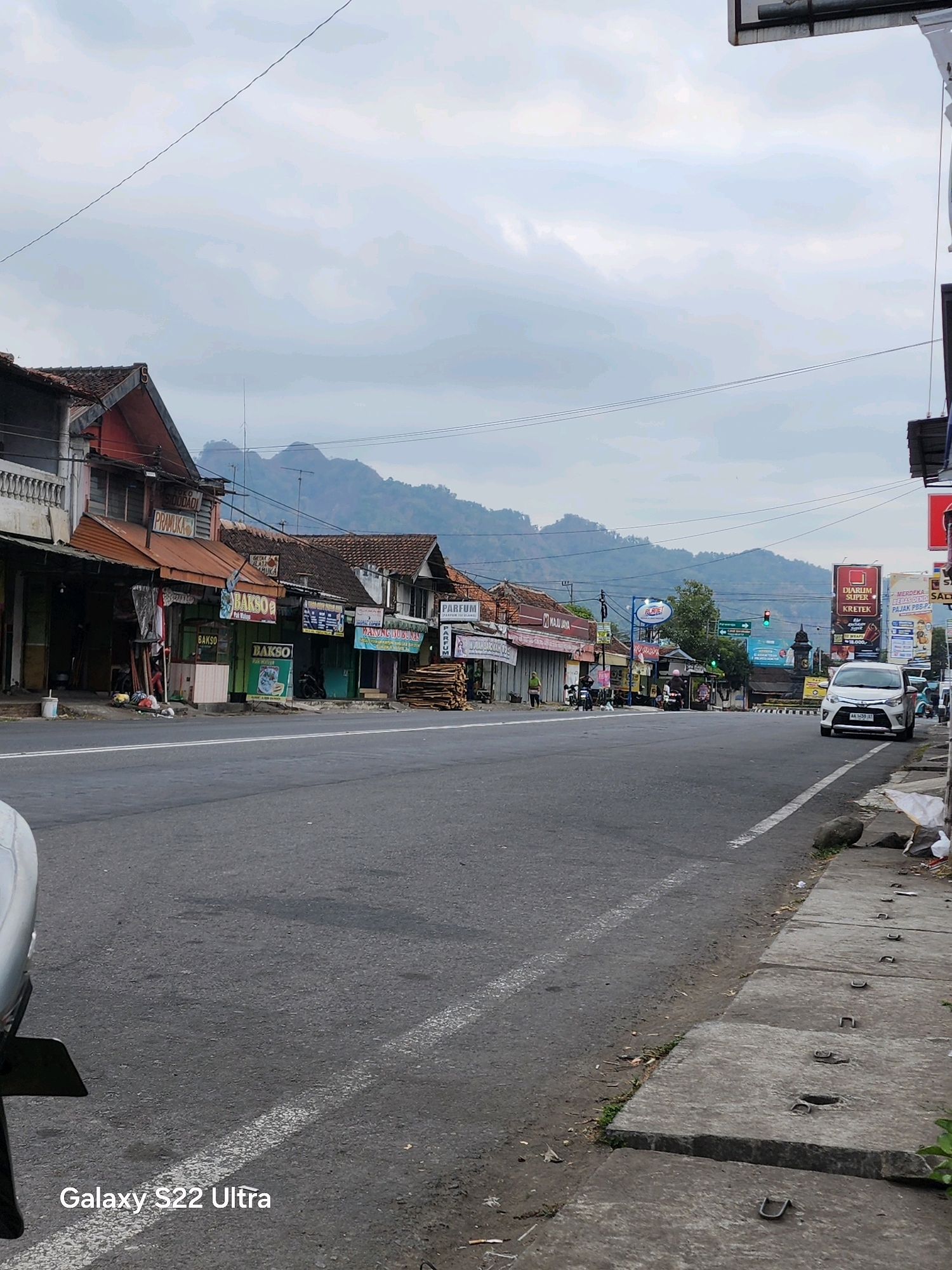 A wide, paved road runs through a town with buildings lining the left side. Mountains are visible in the distance under a cloudy sky. The buildings on the left appear to be shops and businesses with awnings and signs, some of which are in Indonesian. A white car is parked on the right side of the road near a sidewalk with concrete slabs. Several motorbikes are also present, some parked and others being ridden. There is some litter on the sidewalk and road. The overall scene depicts a typical street view in a semi-urban or rural area in Indonesia.