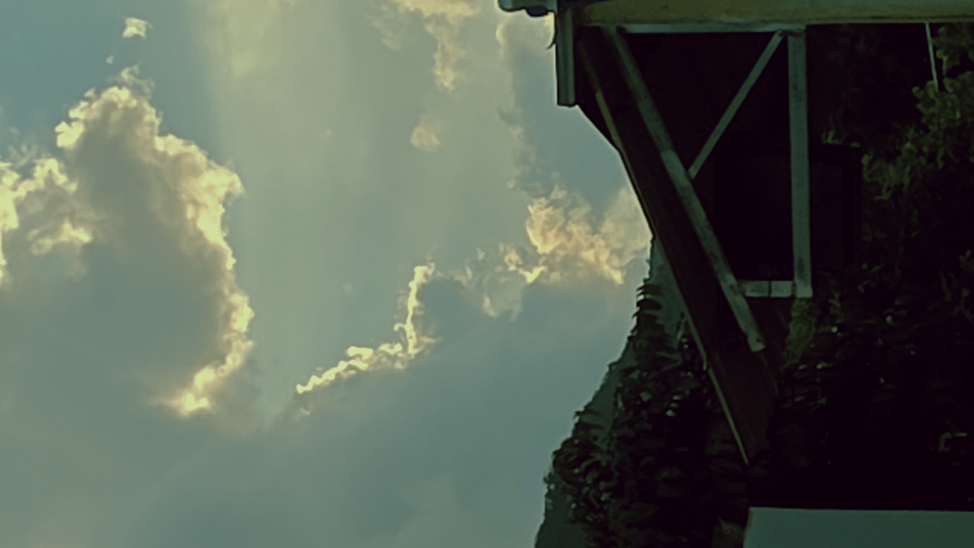 The image is an upward-angled view of a sky with clouds, partially obstructed by the corner of a structure with vegetation. The sky is filled with clouds that have a yellowish-white glow along their edges. A part of a building or shelter is seen on the right side, featuring dark green vegetation and timber framing. It is unknown if this is in the city of Bac Nha Trang Ward, Vietnam.