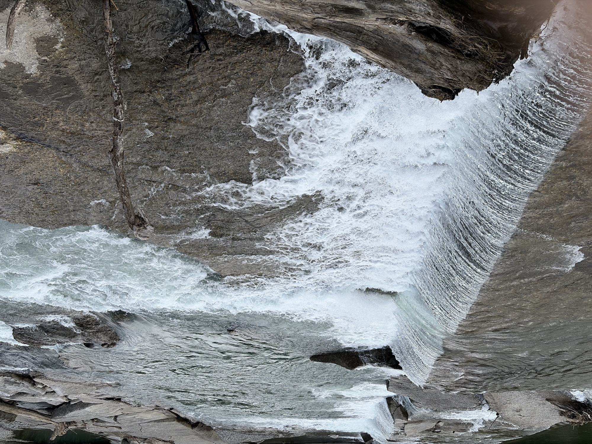 The image shows a fast-flowing river with several small waterfalls and rapids, situated in a natural environment with rocks and driftwood. The water is clear and turbulent, creating white foam and splashes. The riverbed is composed of large, flat rocks and some smaller stones. In the foreground, a large piece of weathered driftwood rests on the bank, its textured surface contrasting with the smooth water. The lighting suggests an overcast day. The image does not contain any people or identifiable structures, and there is no indication of the specific location within the city of Sochi, Russia.