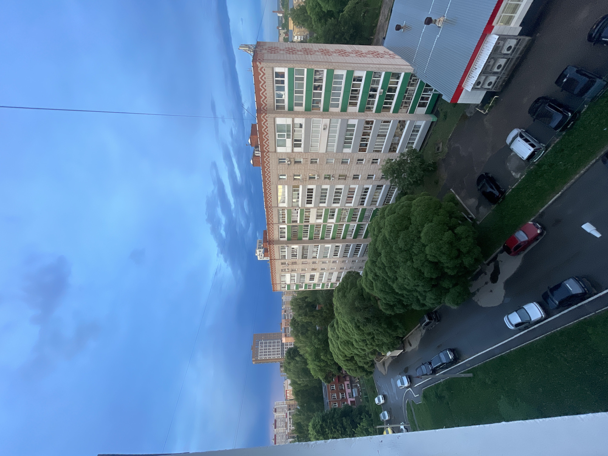 The image depicts a residential scene in Izhevsk, Russia, taken from an elevated perspective. The sky is blue with striated clouds. A high-rise apartment building with green and white accented balconies dominates the midground. Adjacent to it, a lower building with a gray corrugated roof, red trim, and several air conditioning units is visible. Street-level features include lush green trees, a road with parked cars, and a small park or green space with more trees. In the distance are additional buildings. Overhead power lines are strung across the sky.