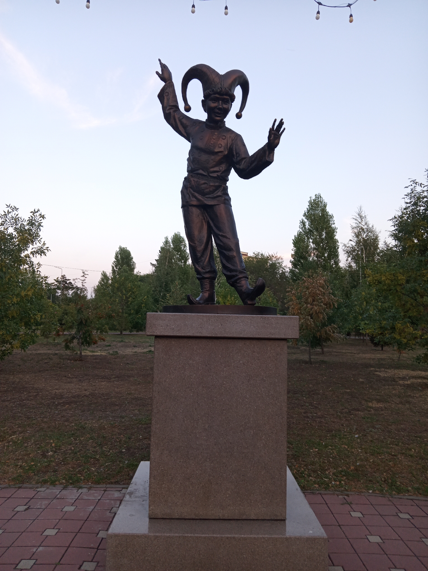 A bronze statue of a jester stands on a rectangular pedestal in a park in Rudny, Kazakhstan. The jester is depicted with a wide smile, a jester's hat with two long, floppy points, and is wearing loose-fitting pants and a tunic. Its arms are raised, with one hand pointing upwards and the other open, as if greeting or performing. The statue is positioned in the center of the frame, with a backdrop of trees and a pale sky. The ground in front of the pedestal is paved with reddish-brown and grey square tiles, and beyond that is a grassy area with fallen leaves. The overall lighting suggests it is late afternoon or early evening.