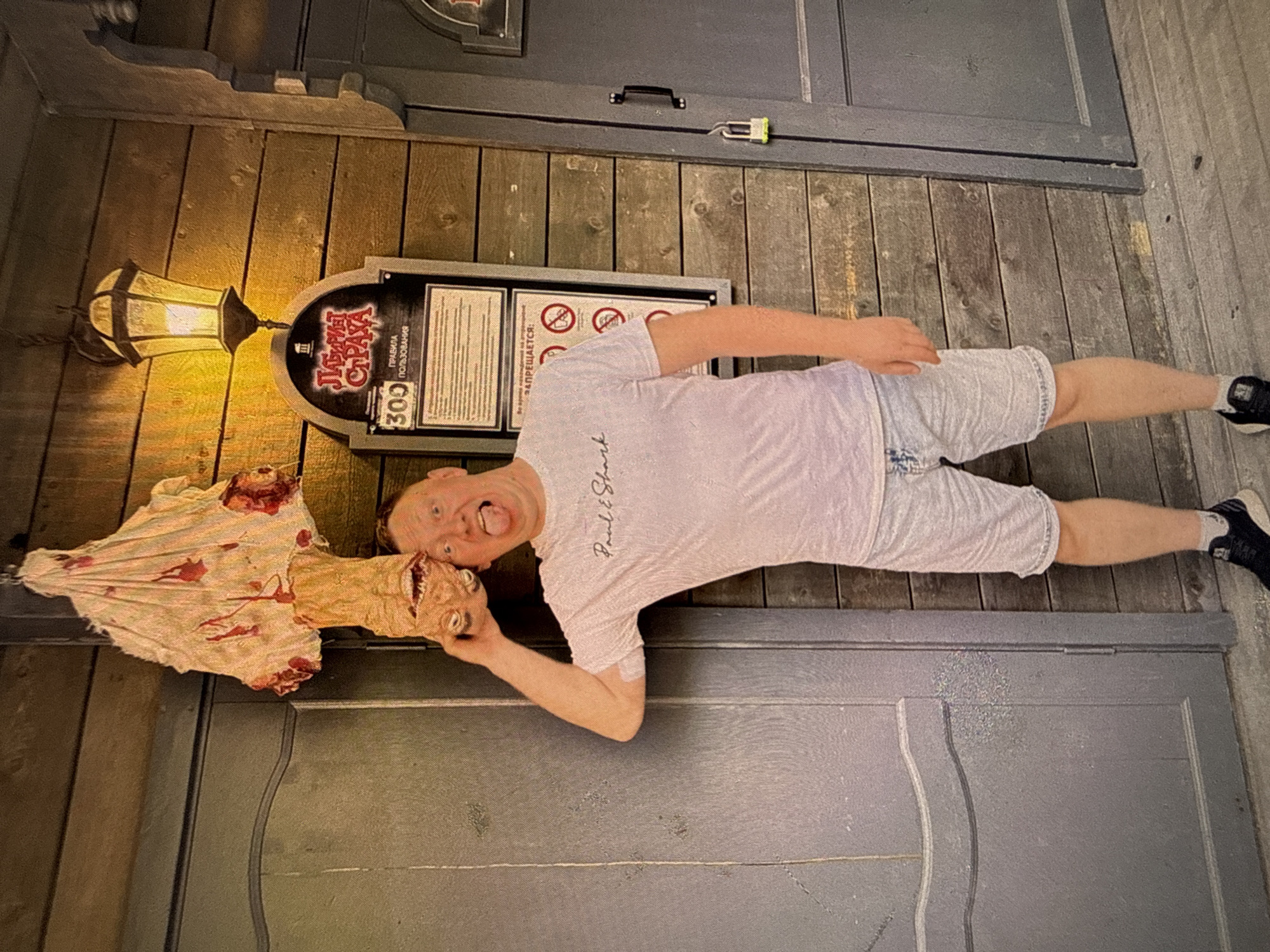 A man stands outside what appears to be an attraction, possibly a haunted house, given the macabre decoration hanging above him. The decoration is a doll or puppet draped in blood-stained fabric, with a grotesque, upside-down head. The man is holding the head of the decoration with his right hand and sticking his tongue out at the camera. He is wearing a light-colored t-shirt with a logo on it, light-colored denim shorts, and dark athletic shoes. Behind him, a wooden wall is visible with a sign that reads "ЛАБИРИНТ СТРАХА" (Labyrinth of Fear) and rules in Russian. A vintage-style lantern hangs above the sign. To the man's left is a dark-colored door, and to his right is another dark-colored door with a padlock on it. The ground is made of wooden planks. The overall scene suggests an outdoor amusement or entertainment venue. The sign and text indicate the location is likely in Chaikovsky, Russia.