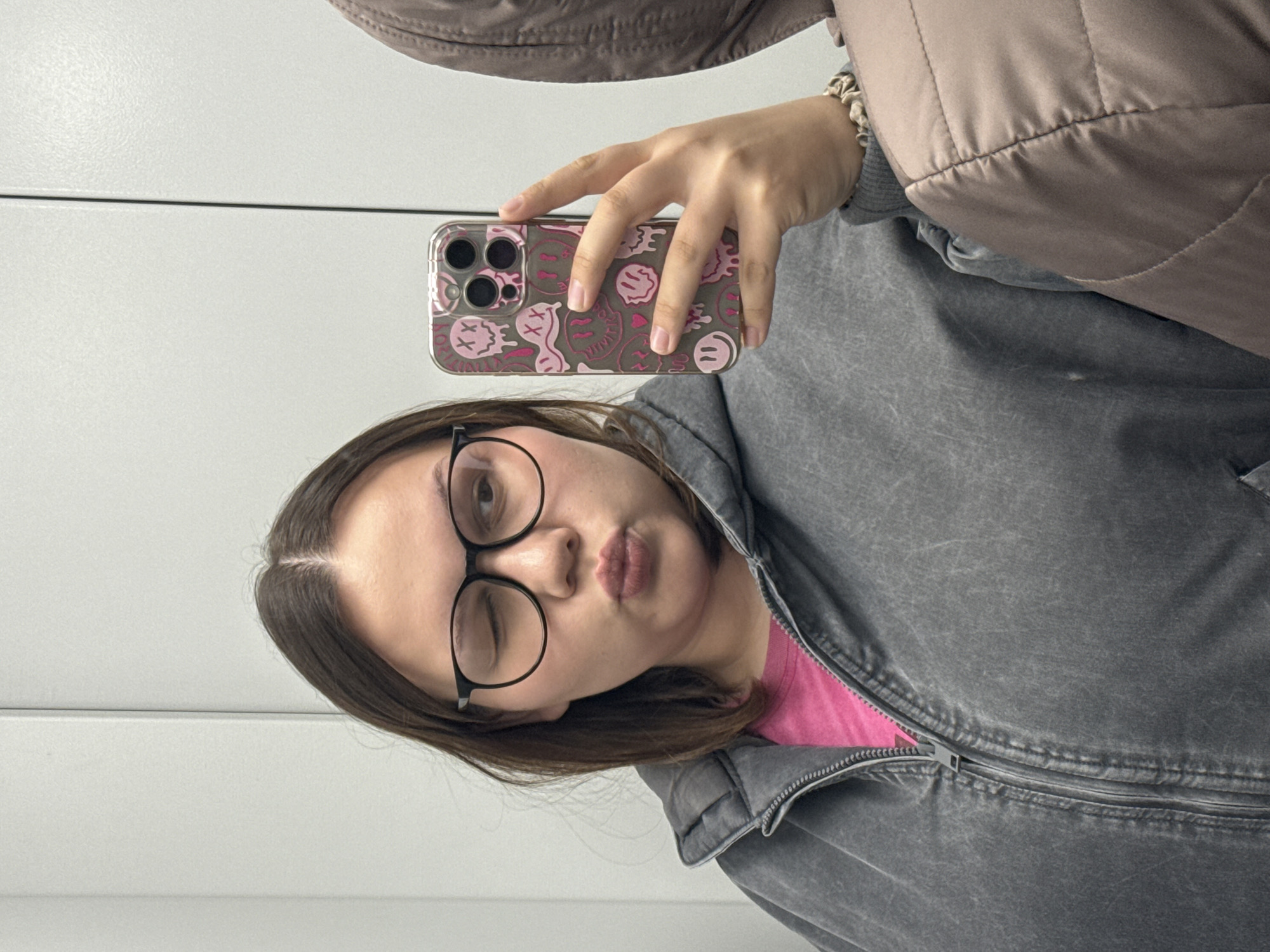 A young woman with brown hair and glasses is taking a selfie. She is wearing a gray jacket over a pink shirt and has a patterned phone case. She is winking and puckering her lips. The background appears to be a white wall. There is no indication of this image's connection to the city of Chaikovsky, Russia.