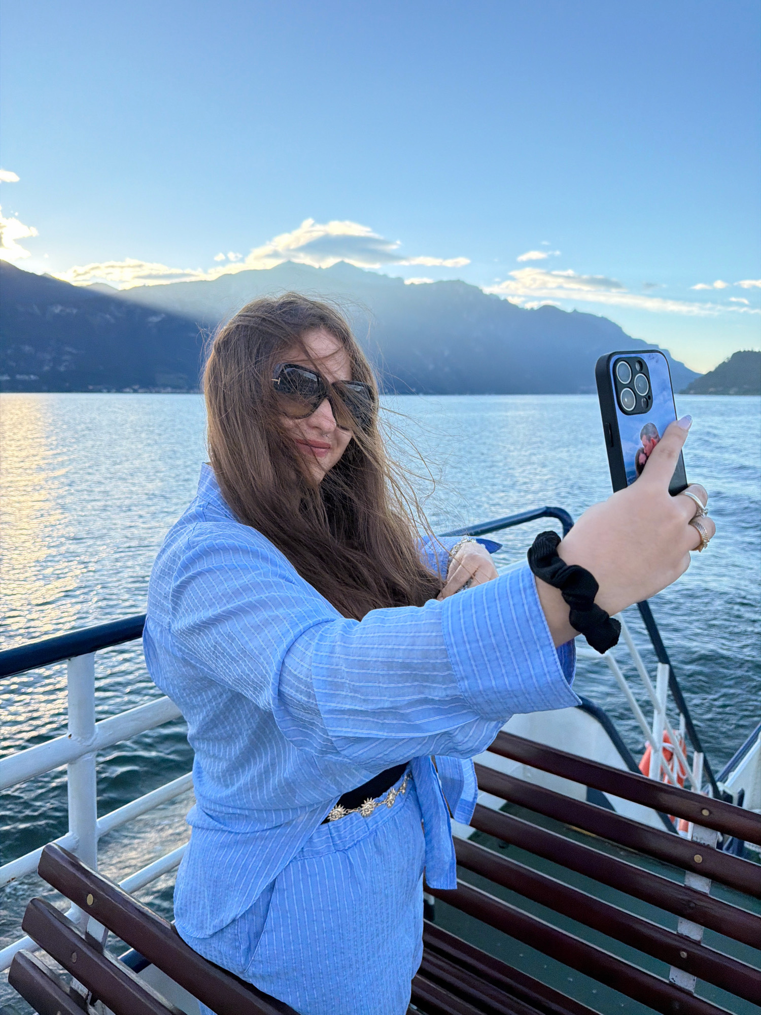 A young woman is taking a selfie on a ferry with a body of water and mountains in the background, likely on Lake Como in the city of Croce, Italy. The woman is wearing a light blue, pinstriped shirt, sunglasses, and has her hair blowing in the wind. She is holding up her phone to take the photo and has a black scrunchie on her wrist. The ferry has wooden benches and white railings. The sky is a clear blue with some clouds and the sun is shining, casting rays over the mountains.