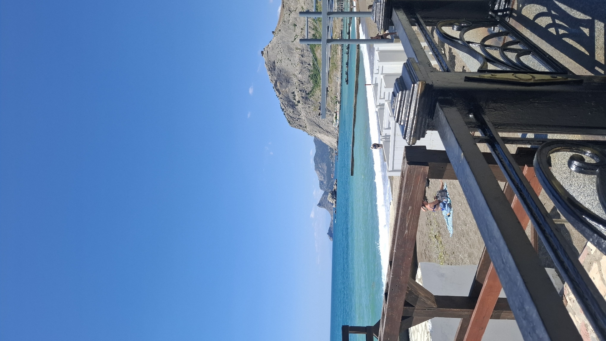 The image depicts a coastal scene with a mountain backdrop and various structural elements in the foreground. A body of turquoise-colored water extends from the foreground towards the base of a mountainous area, which is light grey and sparsely vegetated. The sky above is a clear, cloudless blue.
A series of white, rectangular structures, potentially part of a building or complex, run parallel to the shoreline. In the immediate foreground, a dark metal railing with decorative scrollwork and dark wood support is visible.
On a beach area, which is a light sandy brown color, a person is lying on a blue towel.
The exact location within the city of Unknown, Unknown cannot be determined from the information presented in the image.