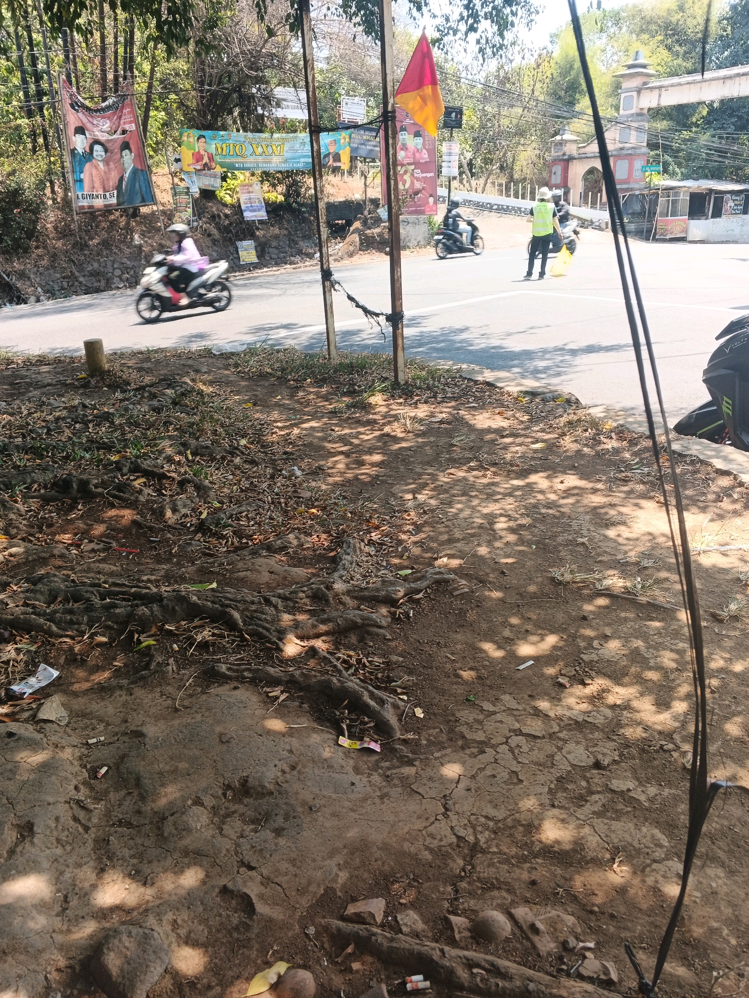 The image depicts a street scene in Semarang, Indonesia, with a focus on the foreground and mid-ground. In the foreground, the ground is covered with dry leaves, exposed tree roots, and scattered debris, including what appears to be cigarette butts. A concrete curb separates the foreground from the road.
In the mid-ground, a road runs horizontally across the image, with a few vehicles. A person on a white motorcycle is moving away from the camera, and another motorcycle is also visible. A person wearing a bright yellow safety vest and a hat is standing in the road, possibly directing traffic. In the background, there are several banners and posters attached to poles, advertising political candidates or events. An archway with decorative architecture is visible in the far background, as is some greenery and trees. The overall impression is a somewhat unkempt urban roadside environment.
