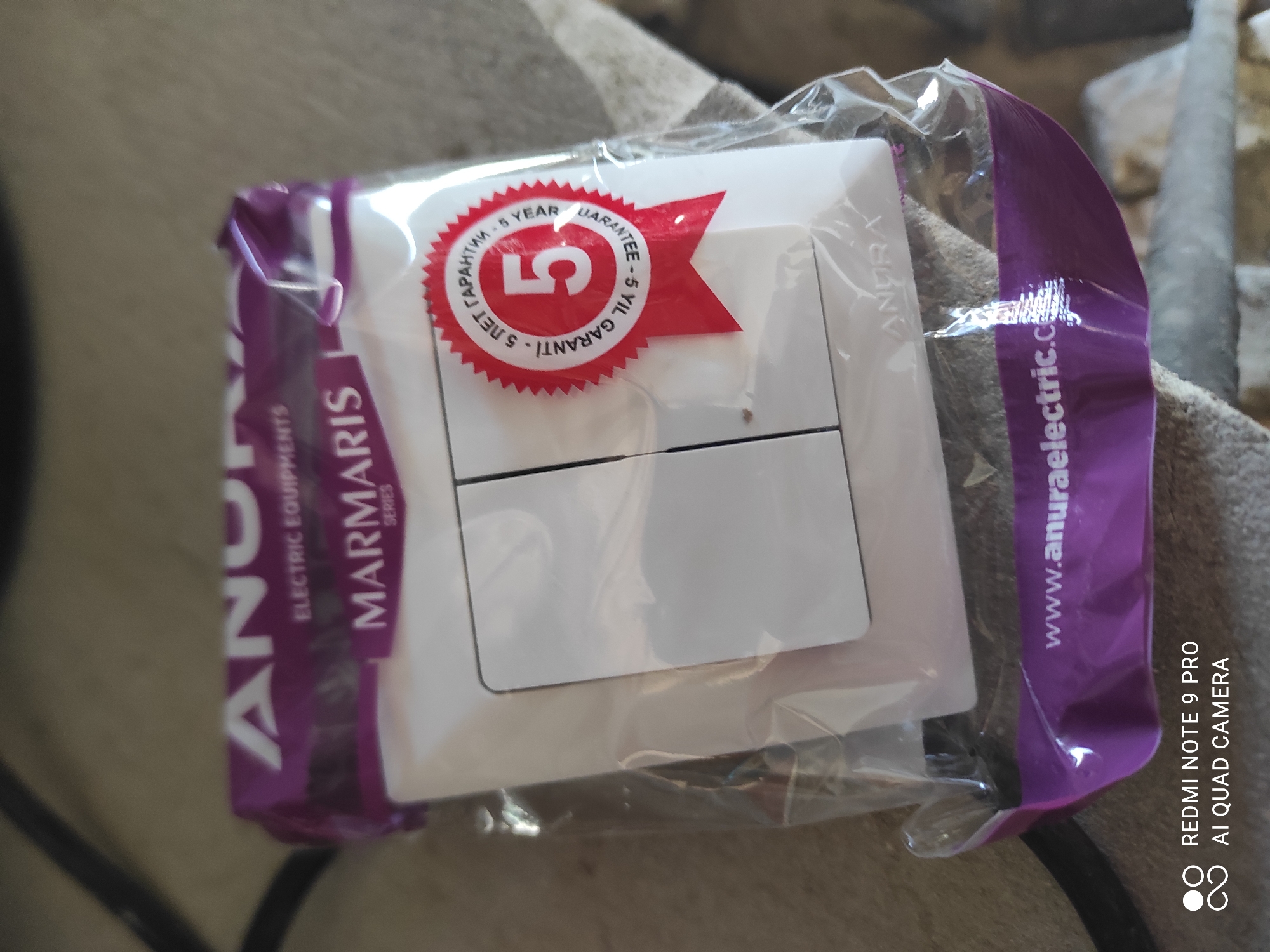 The image displays a close-up, eye-level, indoor shot of a packaged electrical switch. The product is enclosed in clear plastic packaging with a purple band at the bottom and top edges. The purple band at the top features white text that appears to read "ANURA ELECTRIC EQUIPMENTS MARMARIS SERIES". Centered on the packaging is a white electrical switch with two buttons. A prominent red circular sticker with a serrated edge is affixed to the packaging, displaying "5" in large white numerals and text in a foreign language, likely indicating a 5-year warranty. The bottom purple band has the website "www.anuraelectric.com" printed in white. In the bottom left corner, there is a watermark indicating "REDMI NOTE 9 PRO AI QUAD CAMERA". The background is blurred and appears to be an indoor surface with muted, neutral tones, possibly a rug or carpet. The image does not contain any elements that would suggest a location within the city of Khistevarz, Tajikistan.