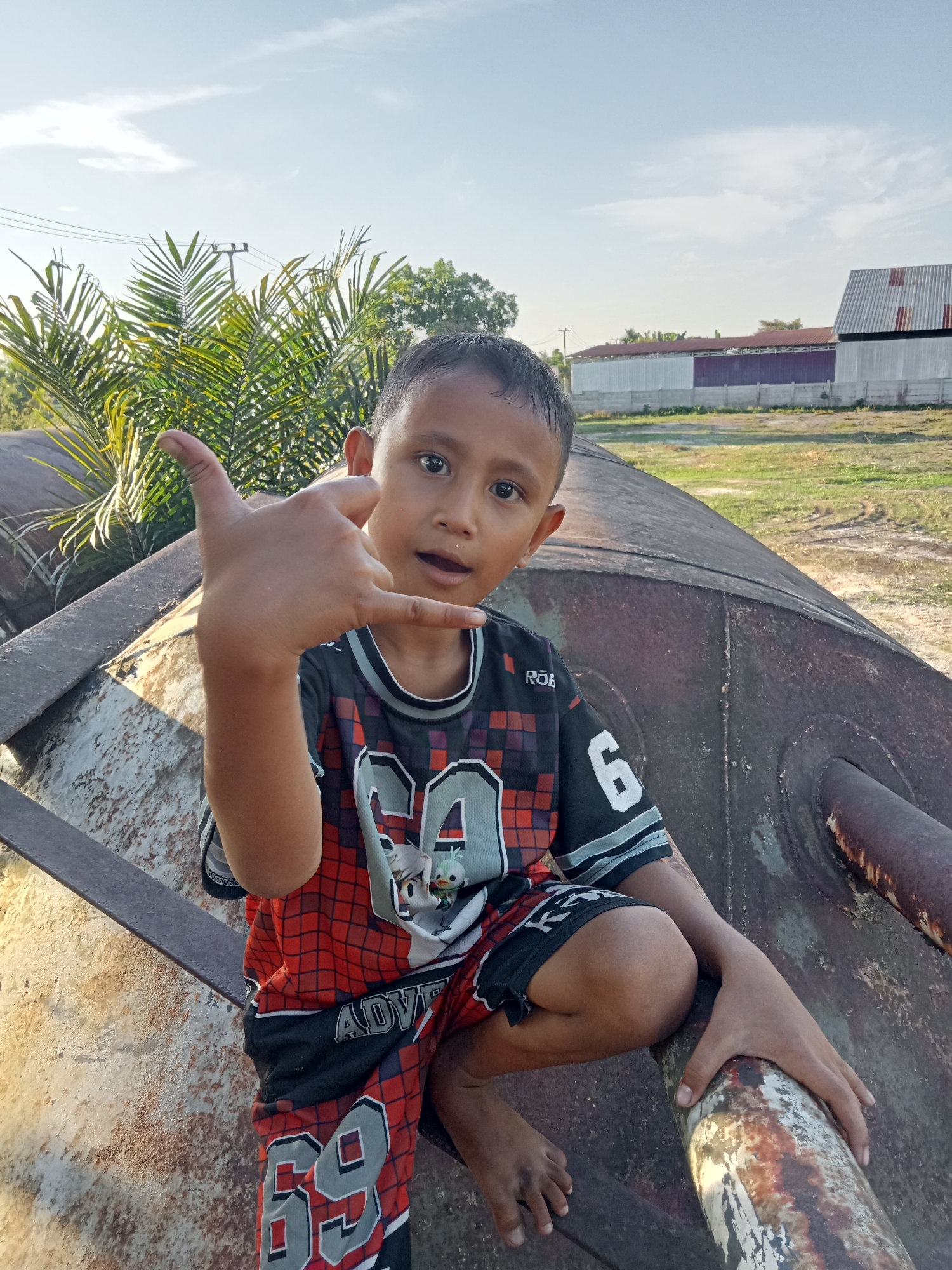 The media file is a photograph of a young boy posing for the camera outdoors. He is squatting on a large, rusty, cylindrical metal object, possibly an industrial pipe or tank. The boy is wearing a black, red, and gray athletic jersey with the number "6" visible on the sleeve and a graphic of what appears to be cartoon characters on the front. He is also wearing black shorts with red and gray accents and the number "69" prominently displayed. His right hand is held up, forming a "shaka" or "hang loose" gesture.
The setting appears to be an open area with dry, grassy ground in the background. Behind the boy, there are tall palm trees with green fronds, and further in the distance, there are several long, low buildings with corrugated metal roofs and walls, suggesting an industrial or agricultural facility. A clear blue sky with some wispy clouds suggests it is daytime. The lighting indicates it is likely late afternoon, with the sun casting long shadows and highlighting the texture of the rusty metal object. The boy has a slight smile and direct eye contact with the camera, conveying a playful and confident demeanor. Visible text on his clothing includes "ROE" on the jersey and "ADVENTURE" on the shorts. The context of Kualu, Indonesia, suggests a tropical or subtropical environment.