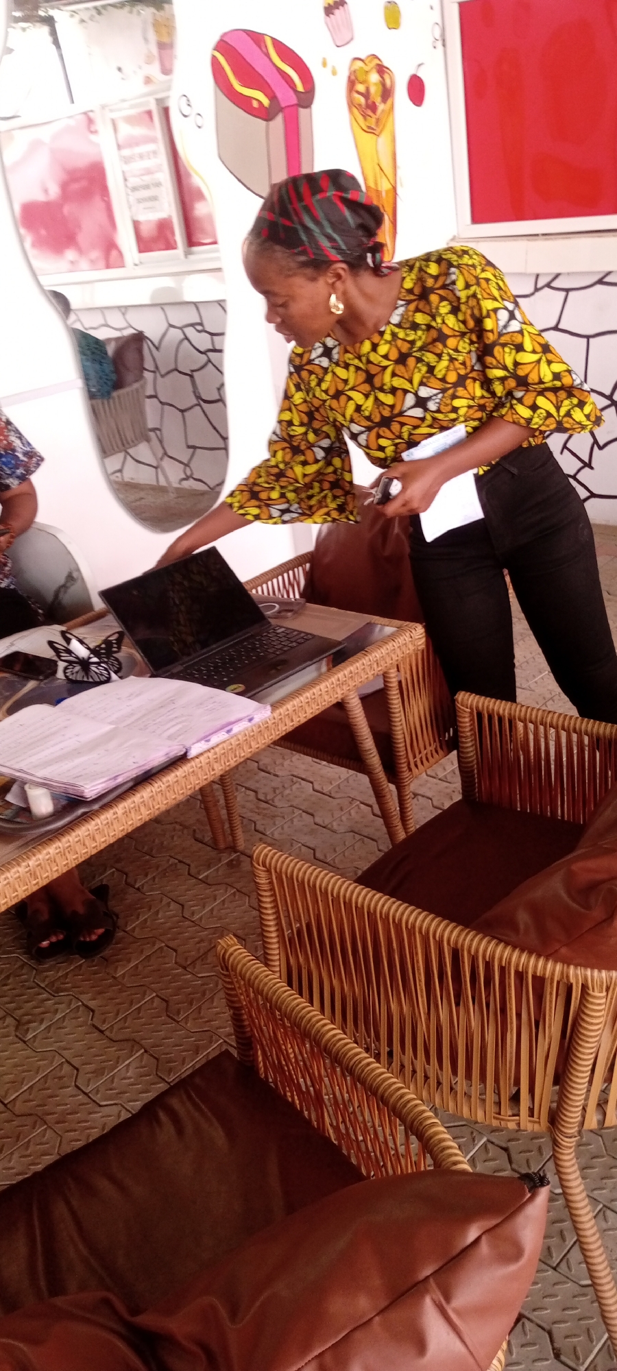 The image depicts a person standing at a table in what appears to be a cafe or eatery in Sabon Gari East, Nigeria, as indicated by the geotag. The individual is a dark-skinned woman with a head covering and earrings, wearing a patterned yellow and brown blouse with sleeves that flare at the ends and black pants. She is holding papers in her left hand and appears to be interacting with a laptop on the table.
The table is made of woven material with a glass top. It holds a laptop, a notebook, and a decorative butterfly paper holder. There are woven chairs with brown cushions around the table. The background shows a wall with what looks like painted or drawn food items, such as a hot dog and a cupcake. There is also a framed window or signage visible in the upper left of the image, with reflective surfaces. The floor is paved with interlocking tiles.