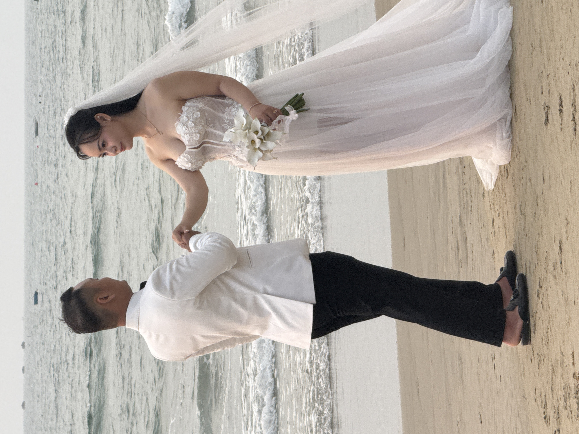 A wedding photo captures a couple on a beach in Đà Nẵng, Vietnam. The bride, wearing a white, strapless wedding dress and veil, holds a bouquet of white flowers. The groom, in a white suit jacket and black pants, holds the bride's hand. They are standing on a sandy beach with waves behind them. The groom is wearing black dress shoes.