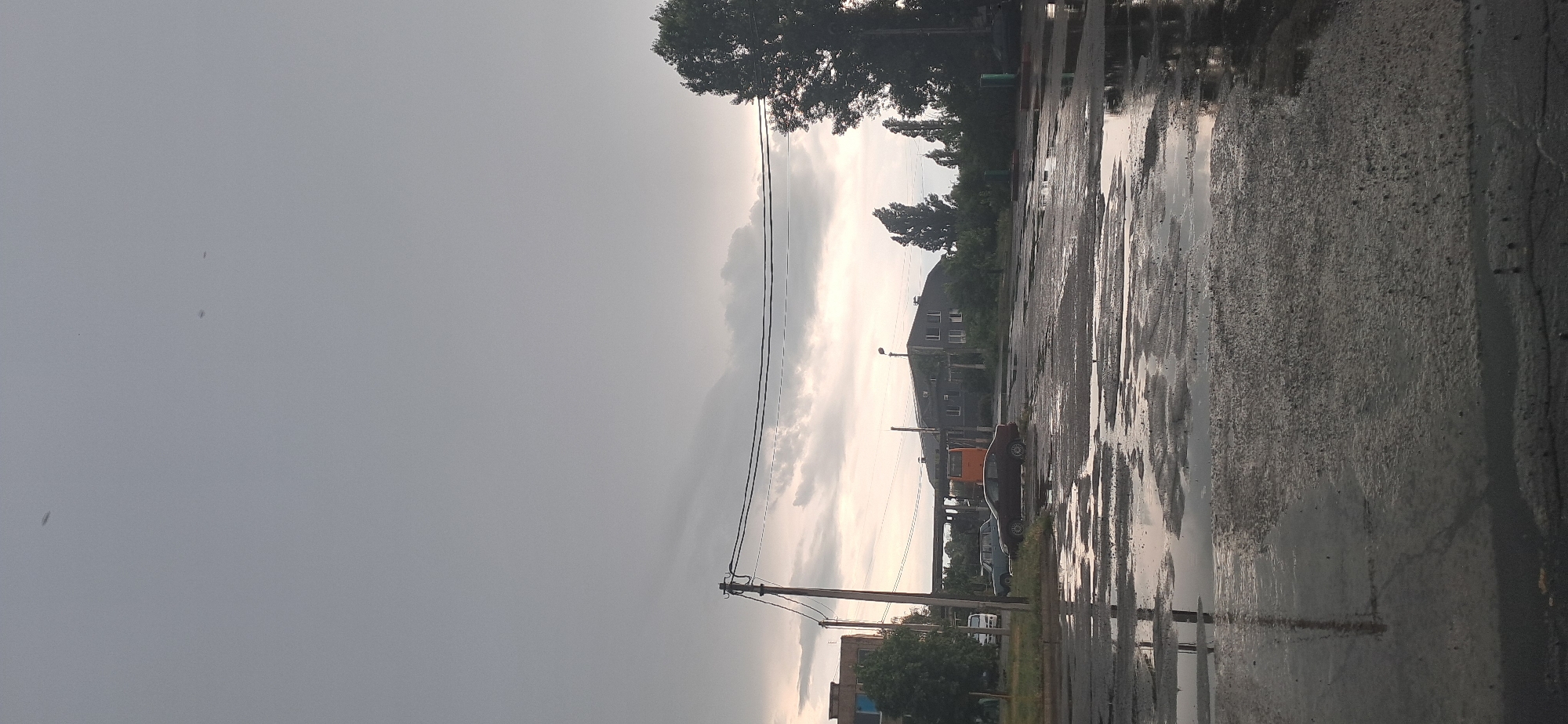 The image depicts a street scene in Horishni Plavni, Ukraine, characterized by overcast skies and a wet, reflective asphalt surface. Several vehicles are present, including a dark sedan parked on the right side of the road and a light-colored minivan further down the street. A public transportation vehicle, possibly a bus, is partially visible near a building complex. The architecture suggests an urban or industrial setting, with multi-story buildings and utility poles visible. The presence of puddles and reflections on the road indicates recent rainfall. The overall atmosphere is subdued due to the heavy cloud cover.
