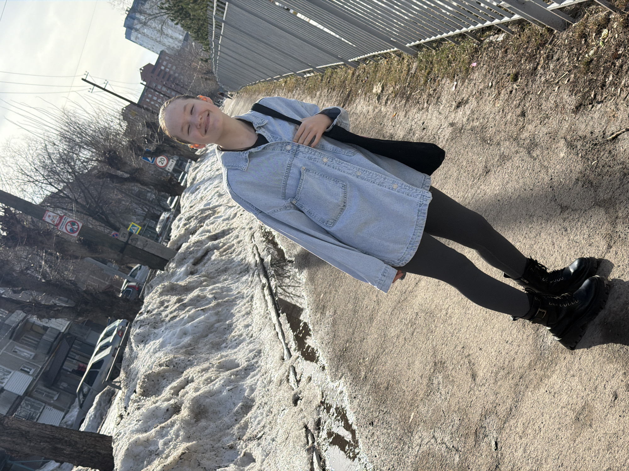 A fair-skinned young adult is standing on a sunlit sidewalk in Perm, Russia. The person is smiling and wearing a denim jacket, dark leggings, and black boots. A black bag is slung over their shoulder. To the left of the person, there's a large mound of melting snow along the roadside. Parked cars are visible further in the background. Buildings line the street, including a multi-story brick structure. A metal fence runs parallel to the sidewalk. A speed limit sign showing '20' is visible in the background. Overhead power lines are present. The scene conveys a transitional period between winter and spring in an urban setting.