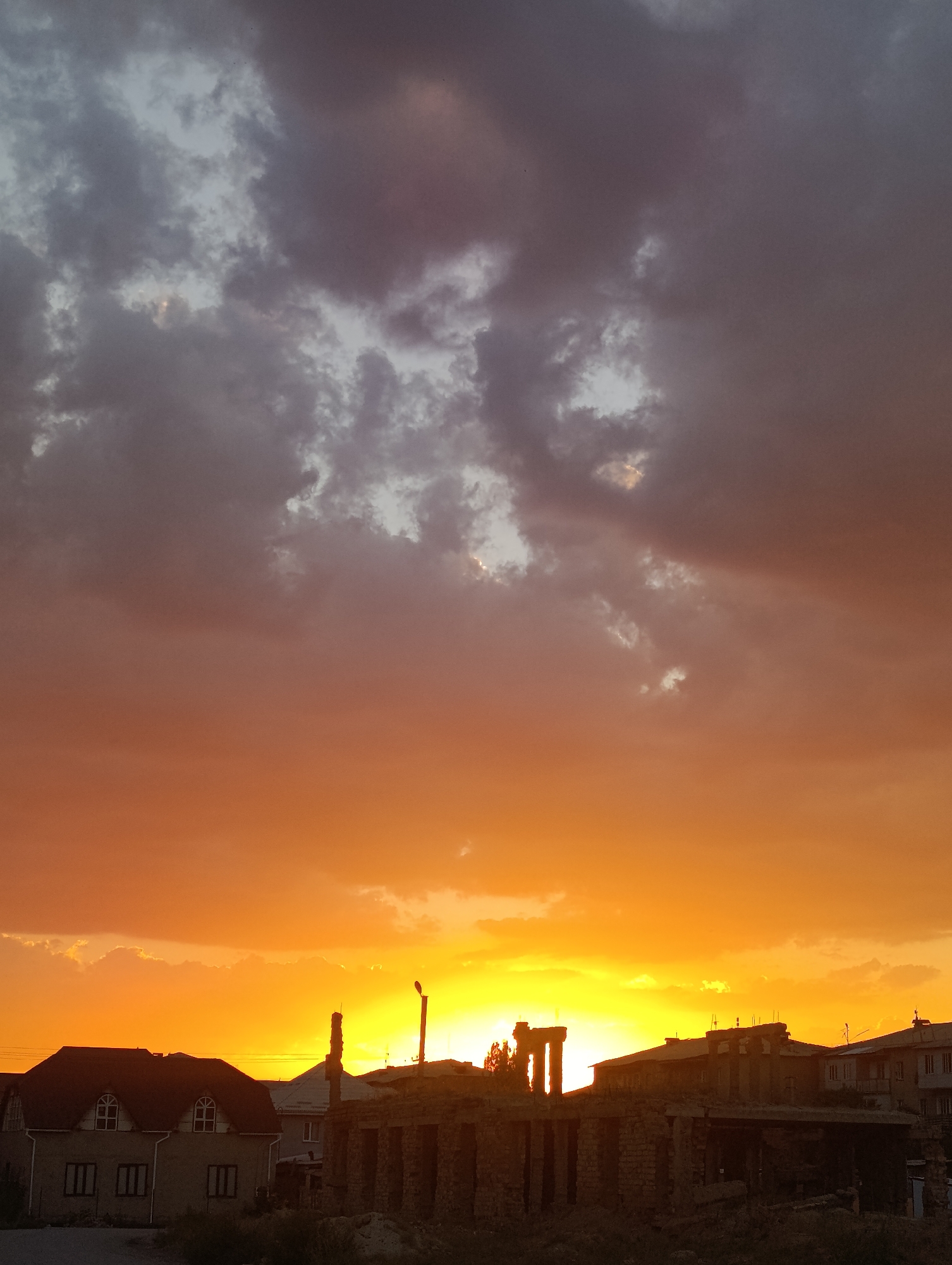 The image depicts a sunset scene in Naryn City, Kyrgyzstan. The sky is a dramatic gradient of orange, yellow, and dusky purple, with scattered dark clouds. In the foreground, silhouetted buildings are visible. On the left, a two-story house with a dark roof and arched dormer windows stands partially obscured by foliage. To its right, a partly constructed brick building with visible doorways and columns dominates the mid-ground. Further to the right, other buildings, including one with multiple windows, are visible, along with a lamppost. The overall atmosphere is one of evening settling over a developing urban landscape.
