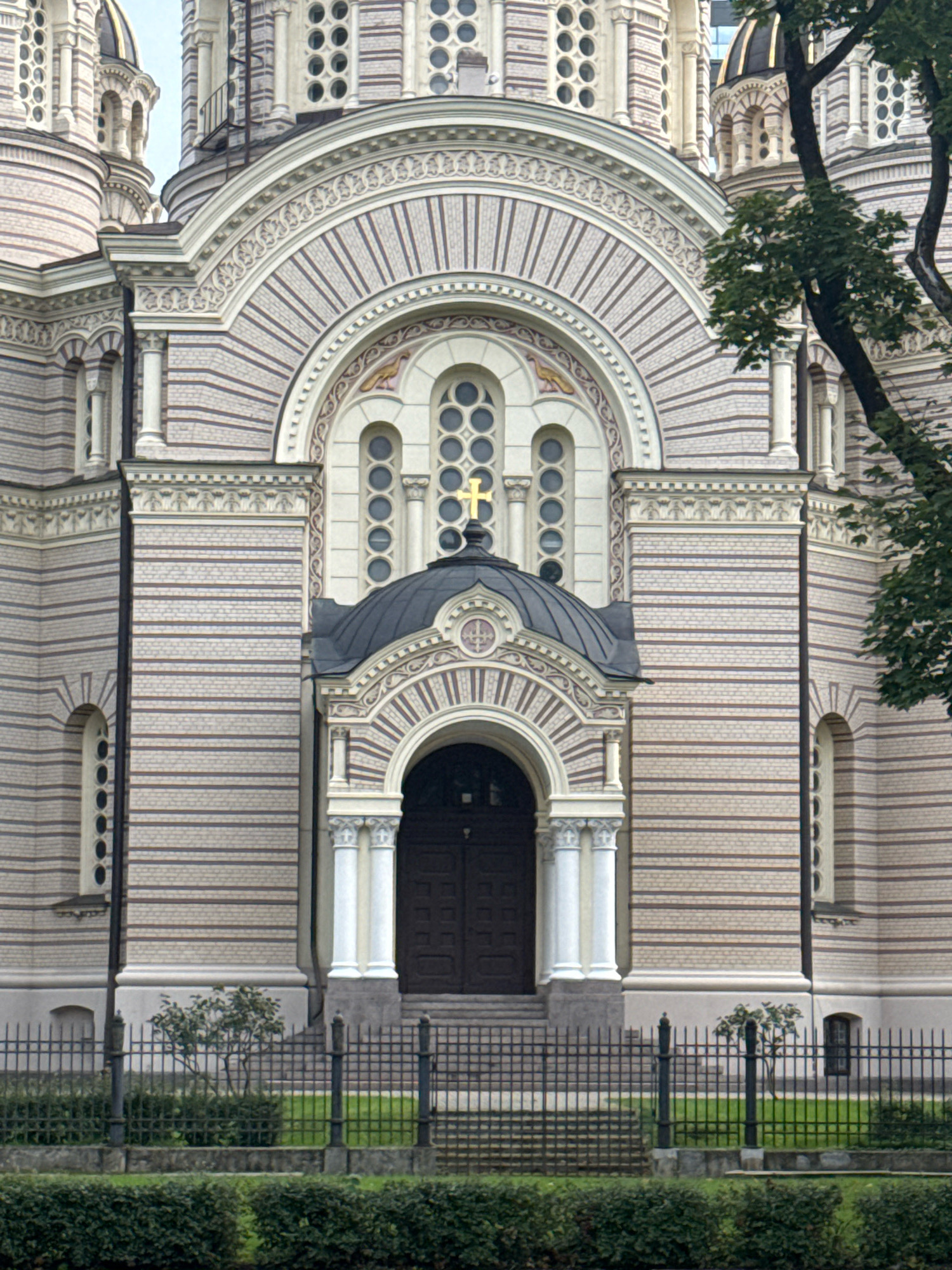 This is a frontal, eye-level, medium shot of the main entrance of the Nativity of Christ Cathedral in Riga, Latvia, on a cloudy day. The cathedral, an Eastern Orthodox church, is constructed with light-colored bricks and dark horizontal stripes, featuring ornate architectural details. The entrance is an arched doorway with a dark wooden door, flanked by two white columns topped with Corinthian capitals. Above the door, a dark, rounded roof is adorned with a gold cross. Rows of circular windows are visible on the facade, and a large, decorative arch with striped detailing frames the entrance. A black metal fence with a low hedge runs along the bottom of the frame, separating the cathedral from the viewer. Lush green trees, partially obscuring the right side of the building, are also present. No people are visible in the image.
