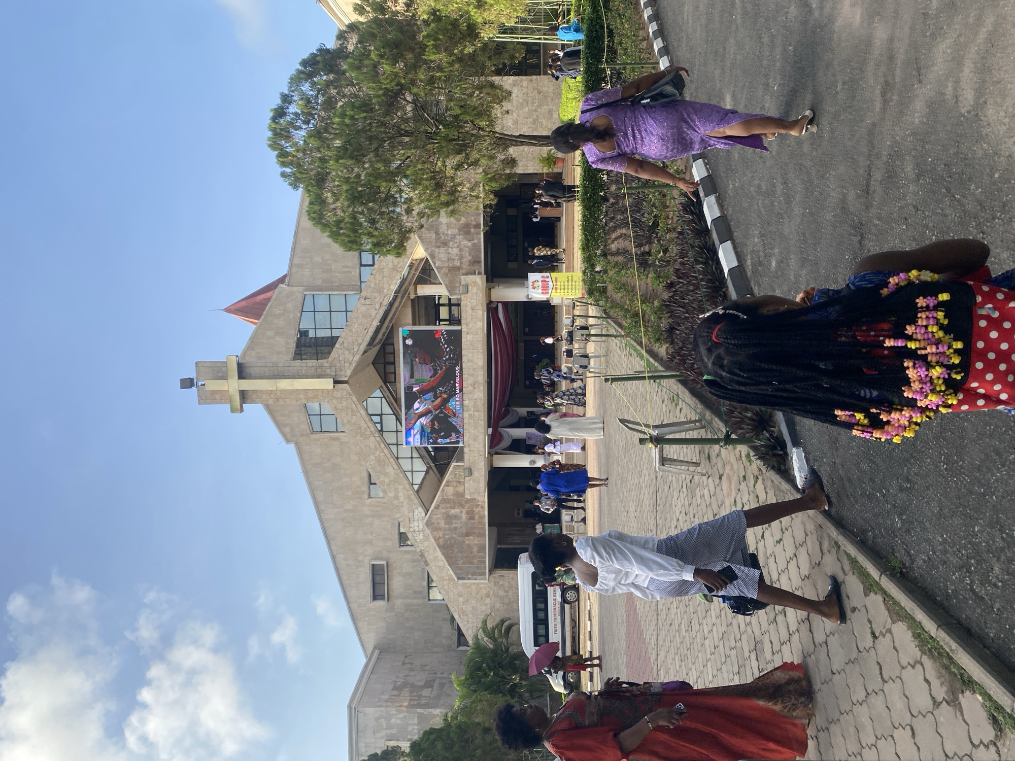 The image shows the Faith Tabernacle Church, Winners Chapel, in Ota, Nigeria. People are walking on a paved path leading to the church building. A banner is displayed on the front of the church. There are a few trees present, and the sky is clear. Some people are wearing purple, white, and red clothing. There is a white van parked in front of the building.