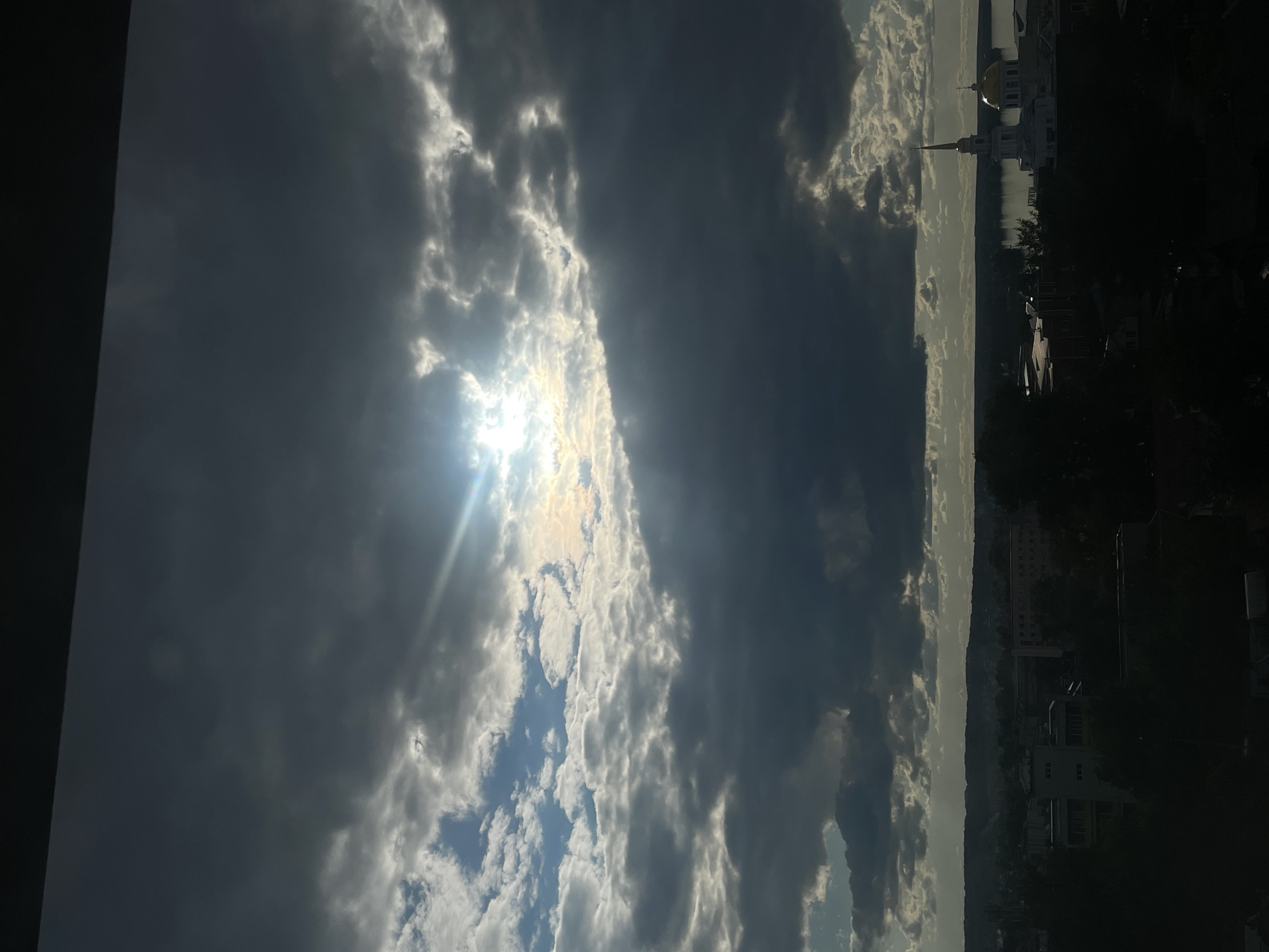 The image captures a dramatic sky with sunlight breaking through thick, dynamic clouds, seen from what appears to be an interior space, with a dark vertical structure framing the far left. The sun is prominent in the upper left, casting intense rays and lens flares through openings in the clouds, creating bright patches of white and light grey. The clouds transition towards the right into darker, heavier grey formations, with glimpses of blue sky visible between them, especially in the lower left.
Along the horizon, a calm body of water, likely the Votkinsk Reservoir, reflects the muted light of the sky. On the distant bank, a cityscape stretches across the mid-right. A notable white architectural complex, featuring a golden dome and a tall, spired steeple, resembling a church or cathedral, stands out against the landscape, consistent with religious architecture found in Votkinsk, Russia. Other buildings and dark foliage are visible in the distance, partially obscured by shadows.
The scene depicts a dynamic weather pattern, likely mid-day to late afternoon, characterized by powerful sunlight piercing through a largely overcast sky. The overall impression is one of natural grandeur and a serene urban-natural blend. There is no visible text in the image.