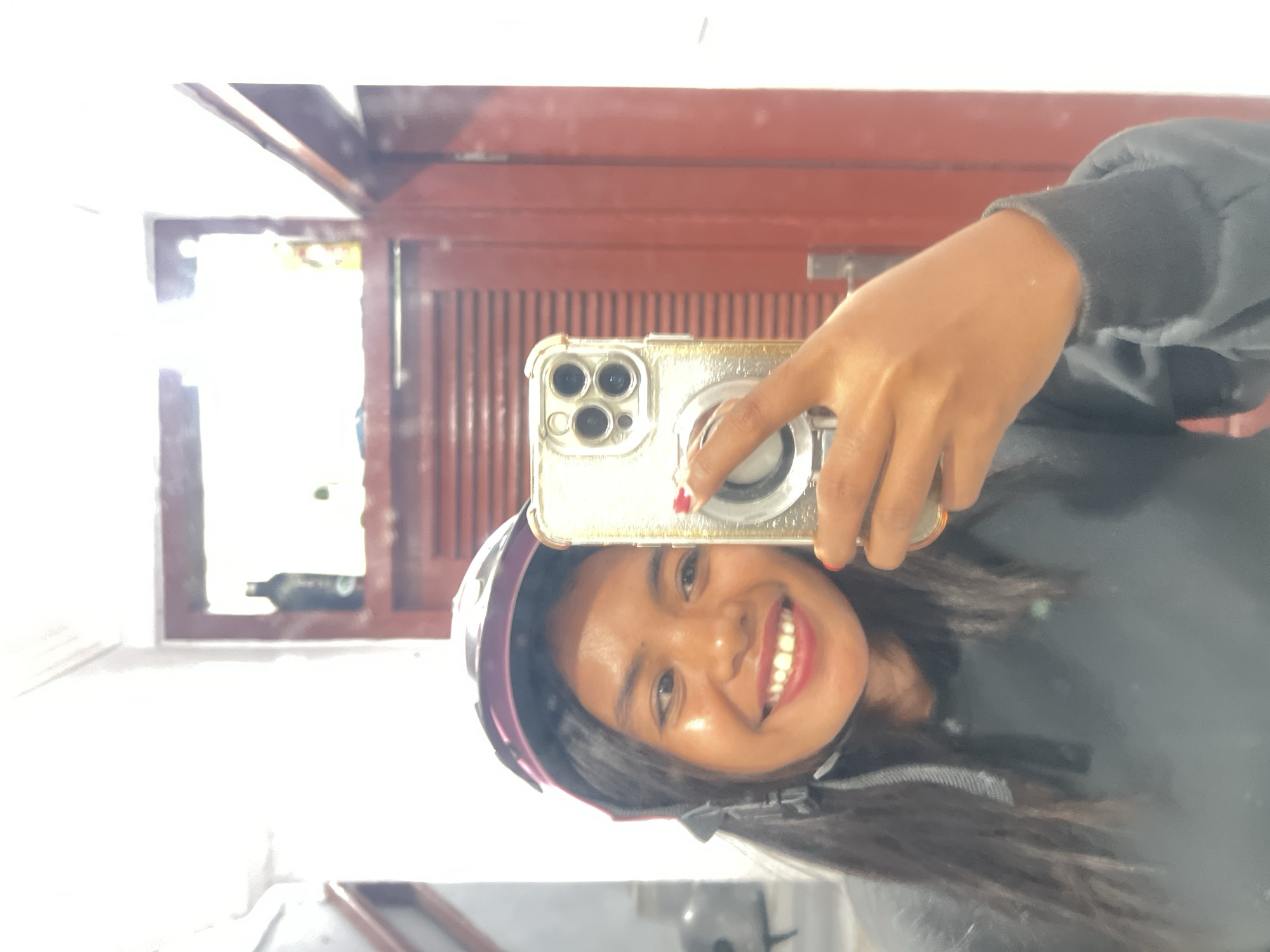A young woman with long, dark hair, wearing a purple and black helmet and a dark grey hoodie, smiles at the camera while taking a selfie with a gold-colored smartphone. Her right hand is holding the phone up to take the picture. The background appears to be a bathroom, with a large mirror reflecting the scene. A dark red door with horizontal slats is visible behind the woman. On a shelf above the door, there are two bottles, one dark and one clear with yellow contents. The overall lighting is bright and somewhat blown out, making it difficult to discern finer details of the environment. The location is identified as Kuta, Indonesia.