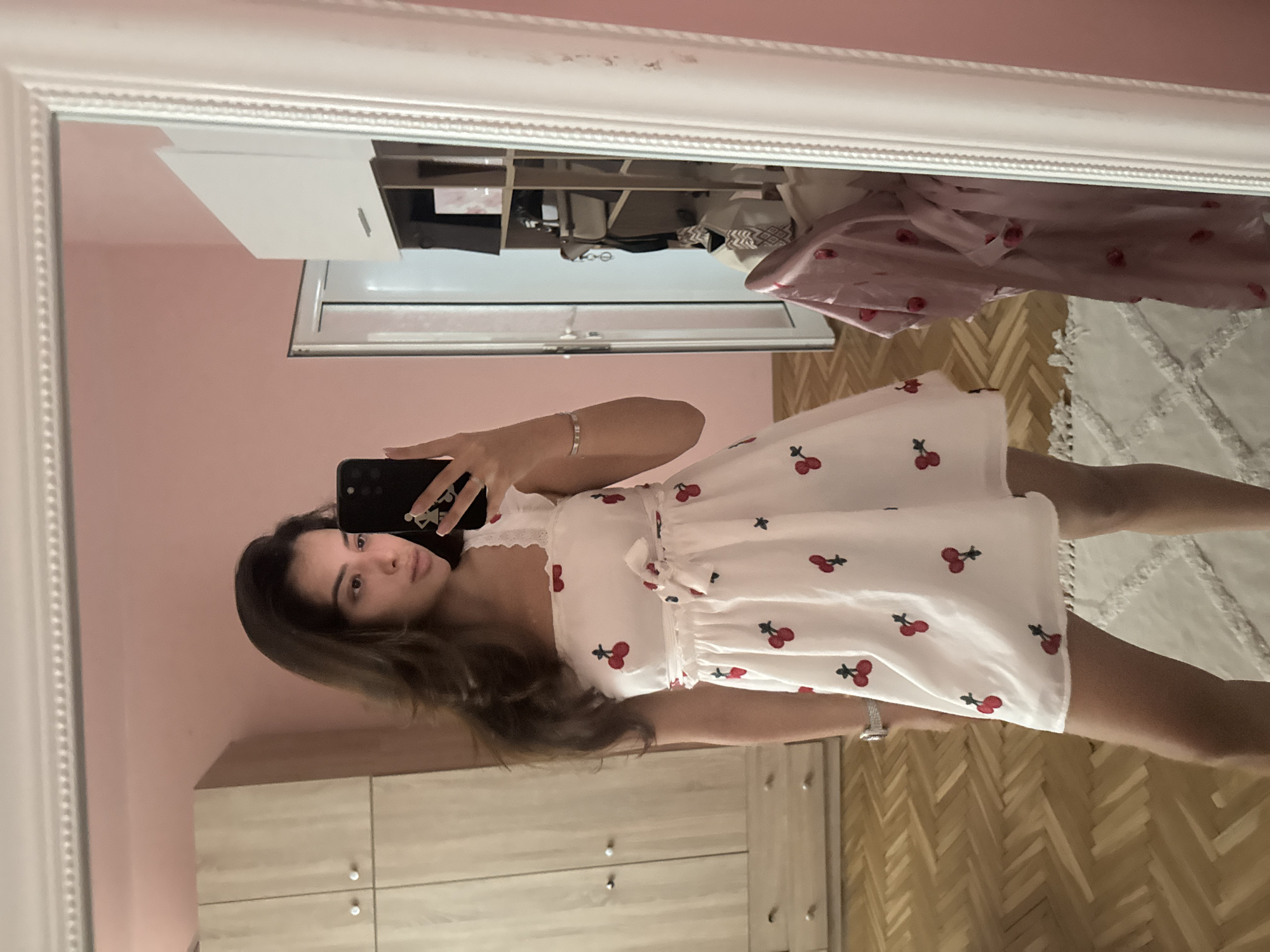 A young woman is taking a mirror selfie in a bedroom in Plovdiv, Bulgaria. She is wearing a white, short, sleeveless dress with a cherry pattern and a bow at the waist. The room has pink walls, a large white-framed mirror, and a wooden floor with a herringbone pattern. A light-colored wardrobe is visible on the left side of the image, and a doorway leads to another room on the right. In the mirror's reflection, a pink garment with a cherry pattern is hanging on a rack.