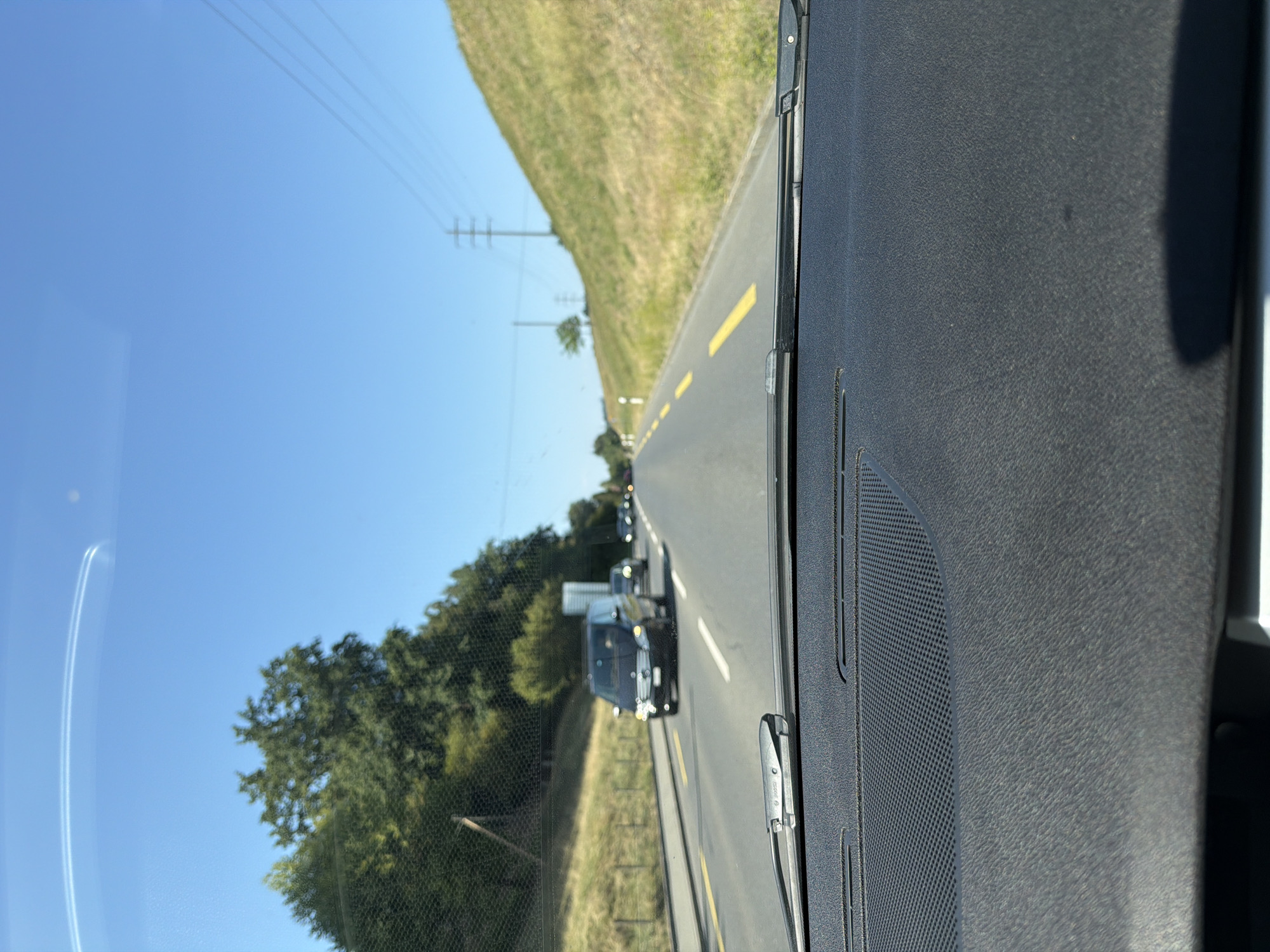 The image is taken from the perspective of someone inside a vehicle driving on a sunny day. The foreground shows the textured dashboard and windshield wipers of the car. The road ahead is paved, with yellow lane markers. A line of cars is visible on the road, including a dark minivan in the middle lane. To the left of the road, there is a grassy embankment lined with trees. To the right of the road, there is a steep, grassy hill with power lines and a utility pole visible in the distance. The sky is clear and blue. The scene is consistent with driving along the Seestrasse in Immensee, Switzerland, a road that runs alongside Lake Zug and features similar scenery.