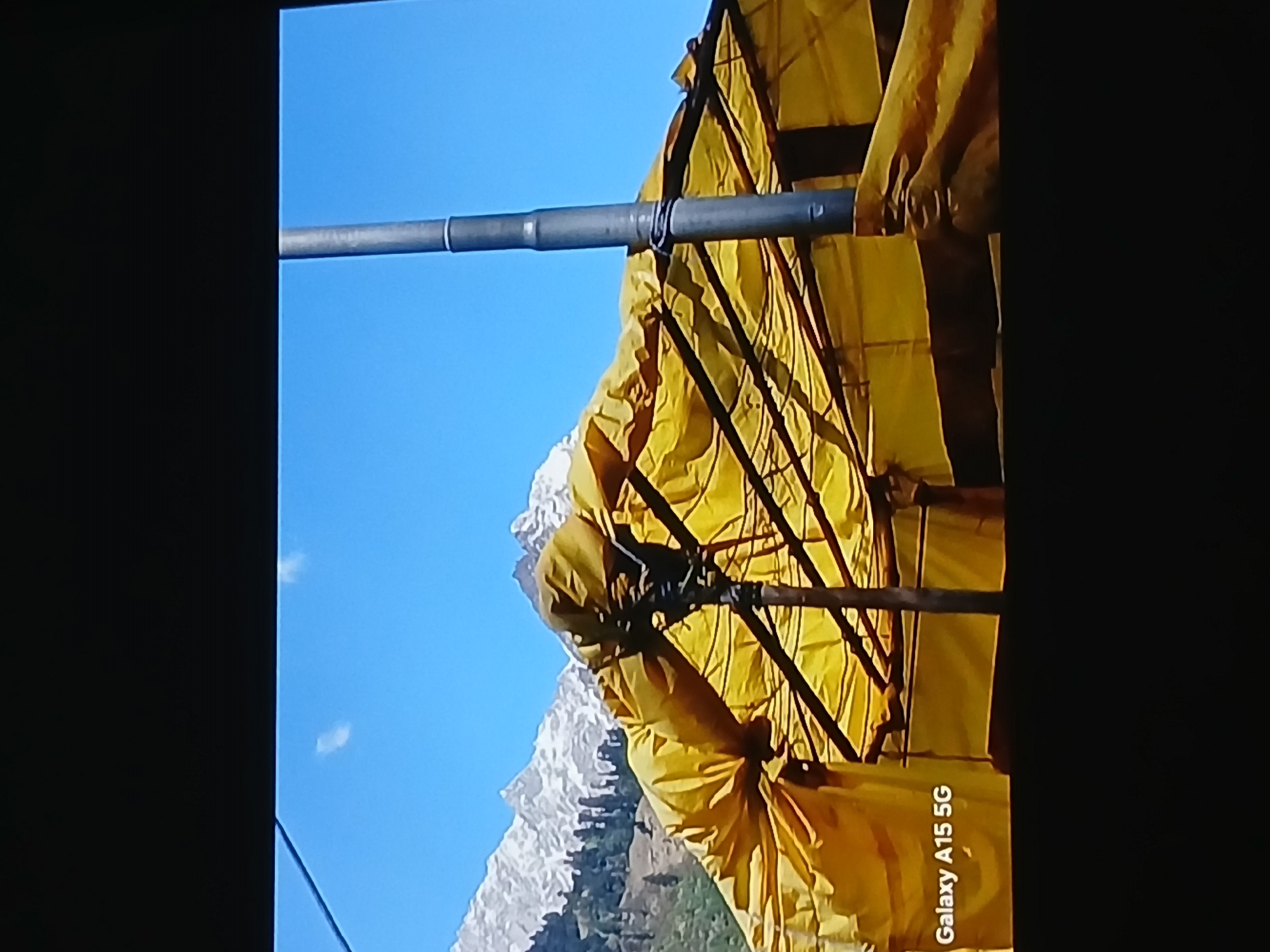 A low-angle shot shows a mountain range under a clear blue sky, partially obscured by a yellow awning supported by a metal frame. The scene is not explicitly identifiable as Bengaluru, India, based on the visual information provided. The composition is framed by black edges on the left and right, suggesting the image was taken from a screen. The words "Galaxy A15 5G" are visible in the lower right corner.