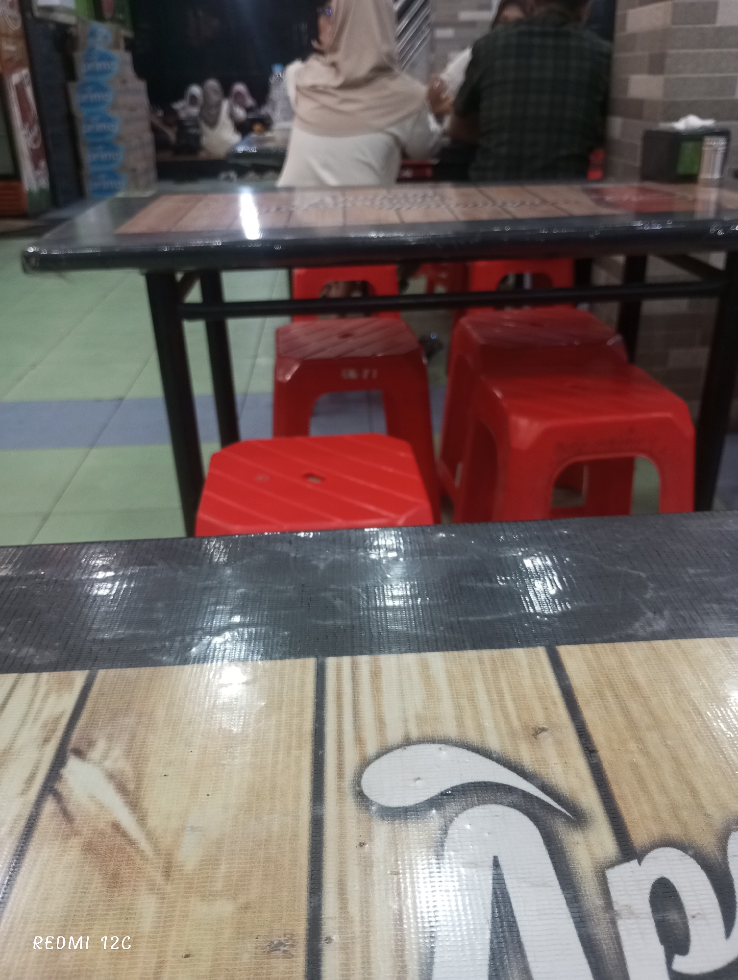 The image shows an indoor scene at an eatery in Lhokseumawe, Indonesia. Several red plastic stools are arranged around tables with a faux wood finish and black trim. Two people are seated at a table in the background; one wears a light-colored headscarf, and the other wears a dark green plaid shirt. Other people are visible indistinctly in the blurred background. In the foreground, a table surface with a wood grain pattern and a partial logo is prominent. The floor appears to be tiled in a light green and blue pattern. The photo was taken using a Redmi 12C phone, indicated by the watermark in the bottom left corner.