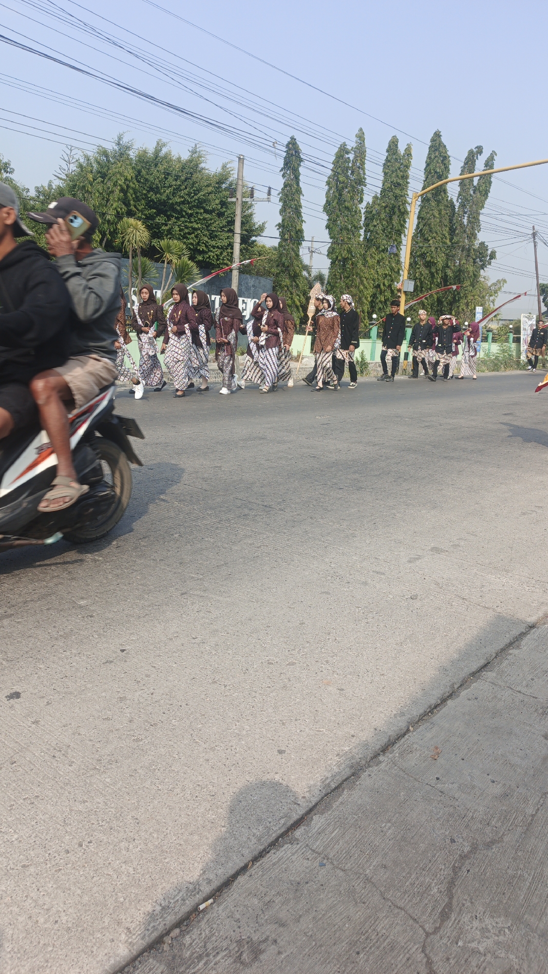 This is a vertical, eye-level, medium shot photograph of a procession in Sumengko, Indonesia. The procession is composed of approximately 15 individuals, all dressed in traditional Indonesian attire. Several women in the foreground are wearing headscarves and patterned fabrics, while men in the background are dressed in dark jackets and patterned pants, some wearing head coverings. The procession is moving from left to right across a paved road. In the lower left quadrant of the image, a person on a motorcycle is partially visible, also moving from left to right. The background features a line of tall, slender trees, power lines, and a yellow traffic signal. The sky is clear and blue, suggesting daytime. The road surface is grey concrete with visible expansion joints and a curb on the right side of the image. The overall scene depicts a cultural event or parade occurring on a street in a village.